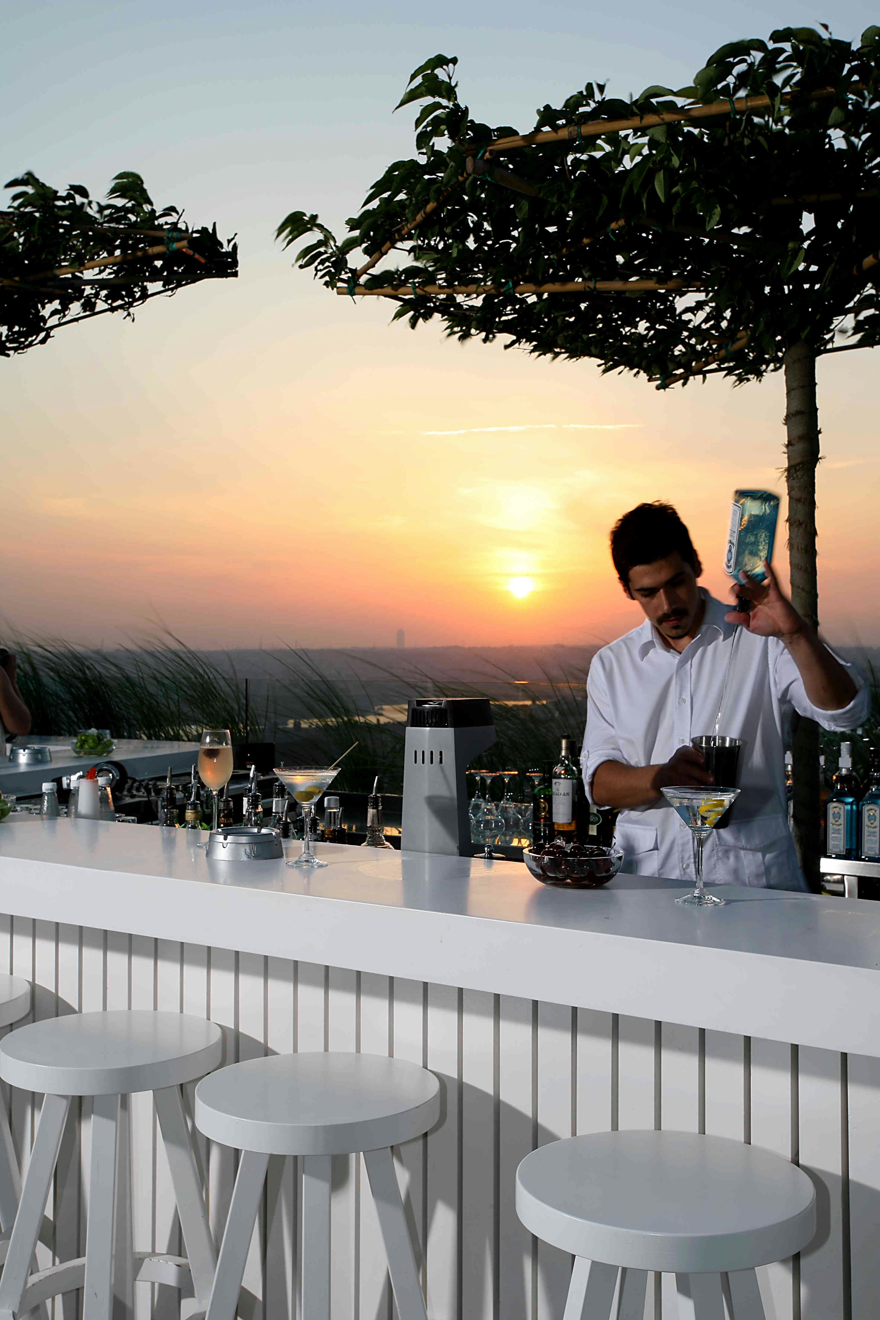 A barman pours gin behind a white bar with four white stools in front of it. The sun sets in the background