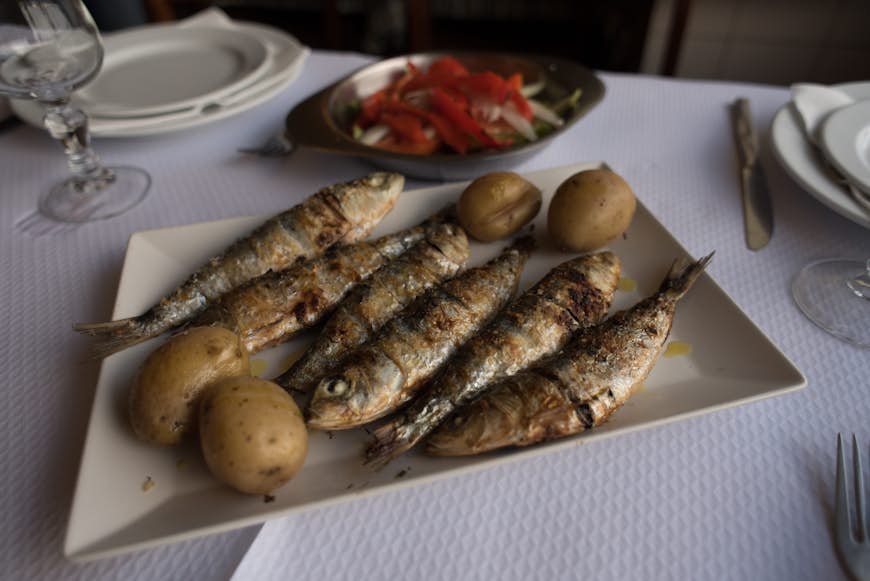 A plate of grilled sardines with baby potatoes and salad on the side, served on a table with a white tablecloth