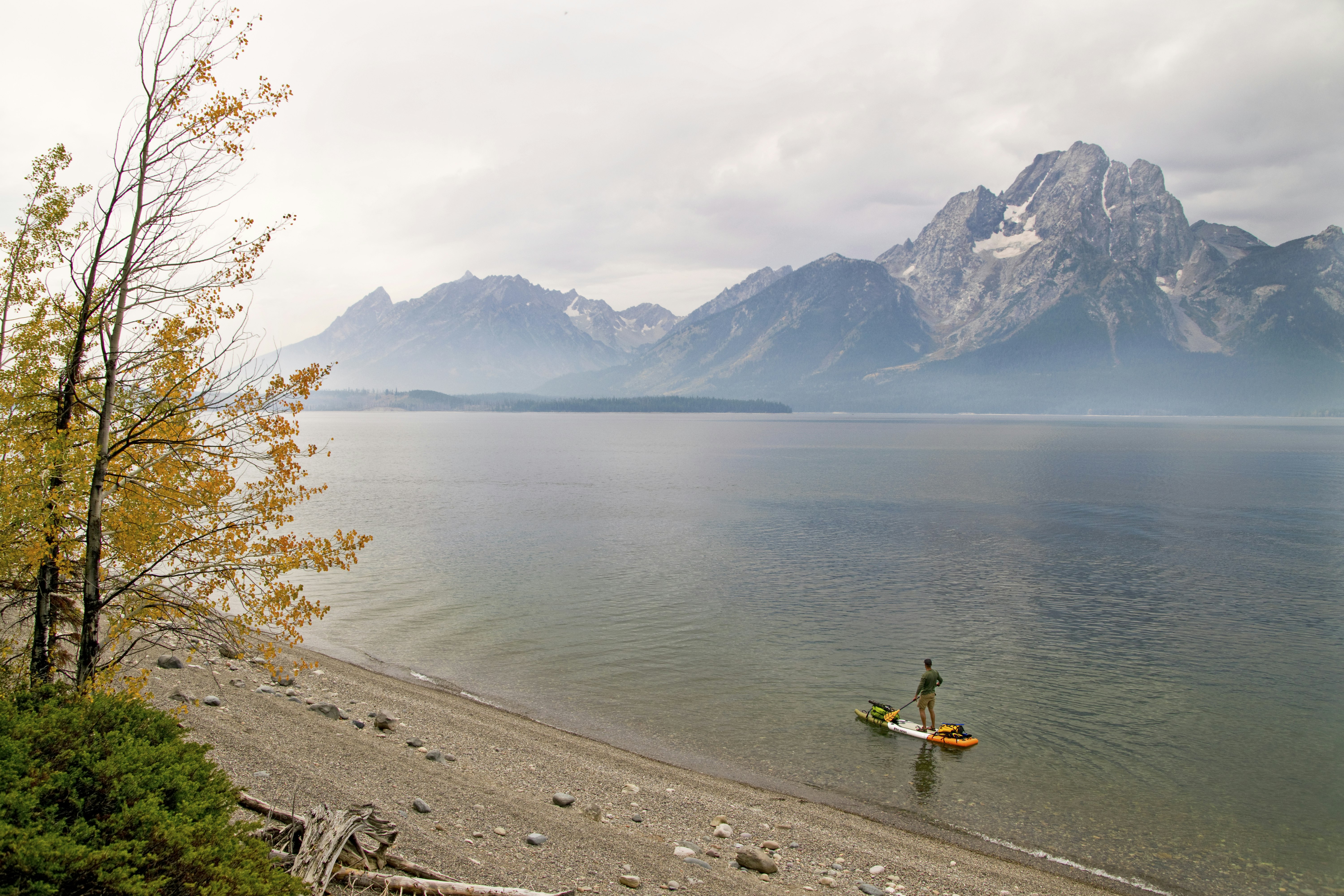 A man paddles a paddleboard across a misty lake with a mountain rising in the background at Grand Teton National Park; Stand up paddle boarding