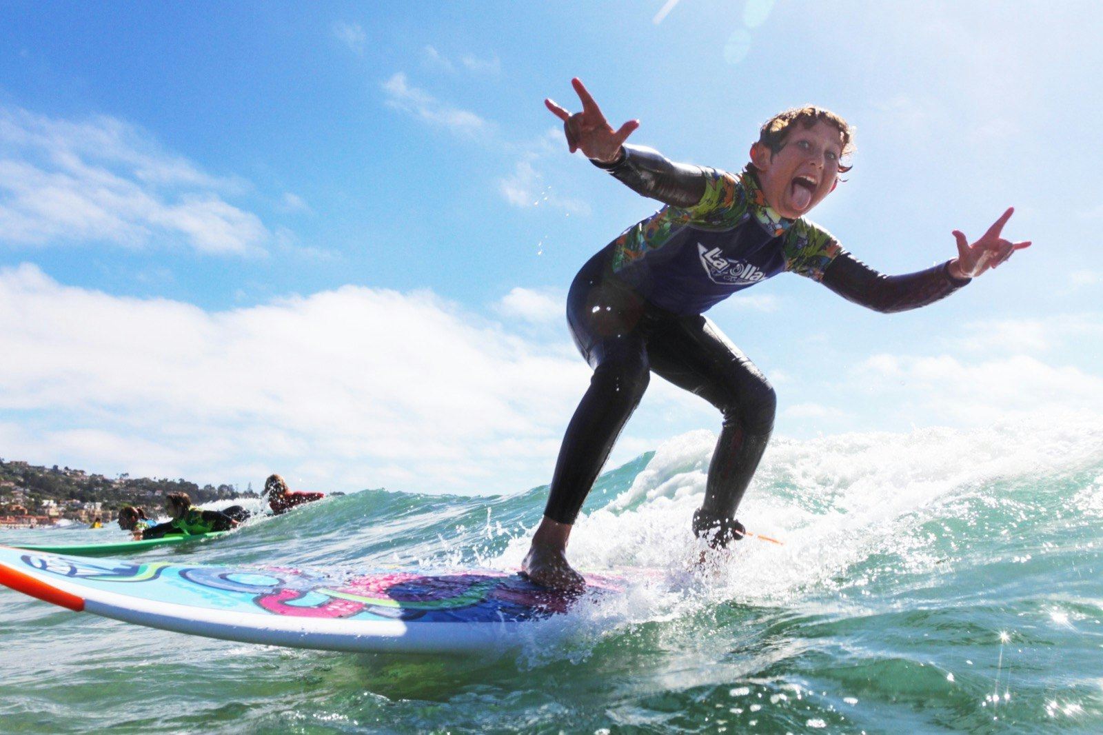 A young boy on a surf board in a wet suit flashes hand signs and makes a funny face while riding a wave