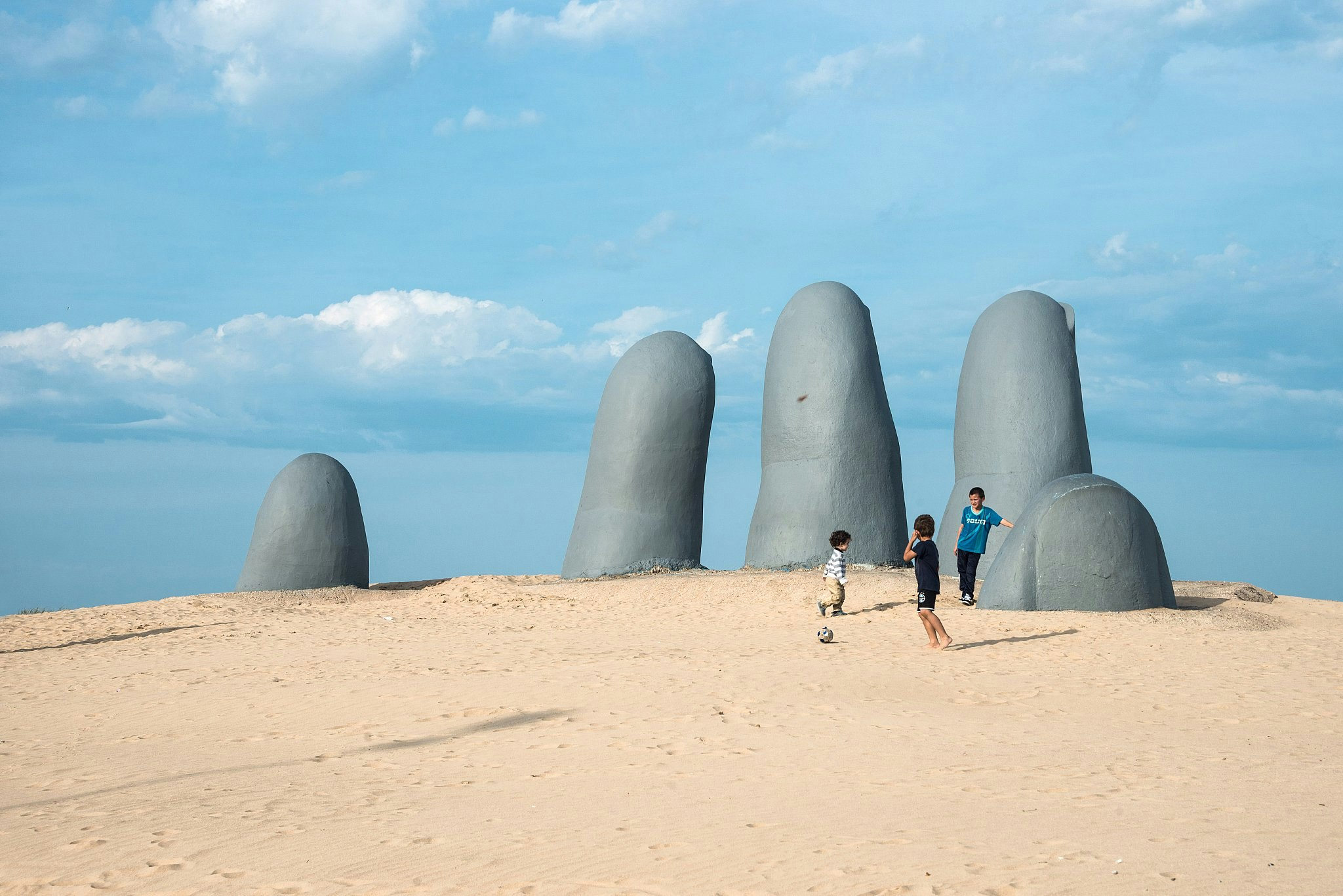 Three boys playing football next to La Mano en la Arena, a sculpture of a huge hand protruding from a sandy beach at Punta del Este, Uruguay.