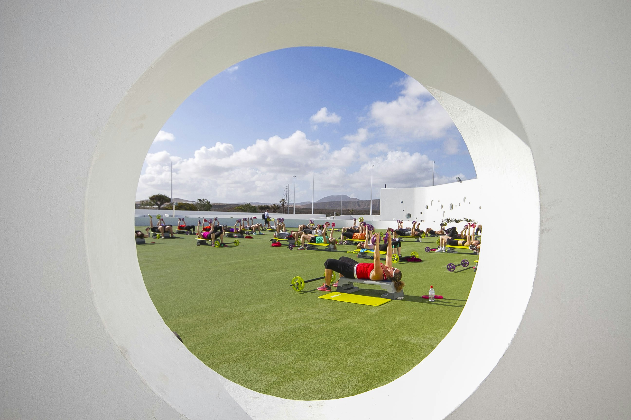 Looking through a circular hole in a pristine white wall, a group of people lay on small benches while performing chest exercises; at their feet are barbells sitting on the artificial grass.