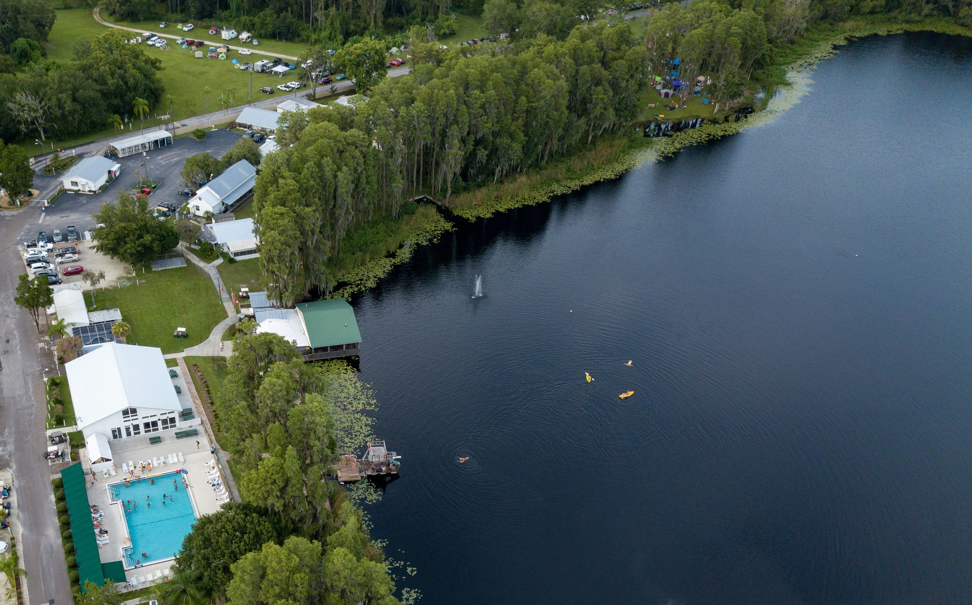 An aerial shot of the Lake Como Resort in Florida. On the left side of the frame, a light blue pool contrasts with the dark blue lake that takes up most of the photo. Tiny peach dots are nude swimmers. The white buildings with metal roofs sprawl out around the lake, the shore lined thickly with green trees. A campground with colorful tents appears amongst the foliage and two orange kayaks at in the middle of the lake.