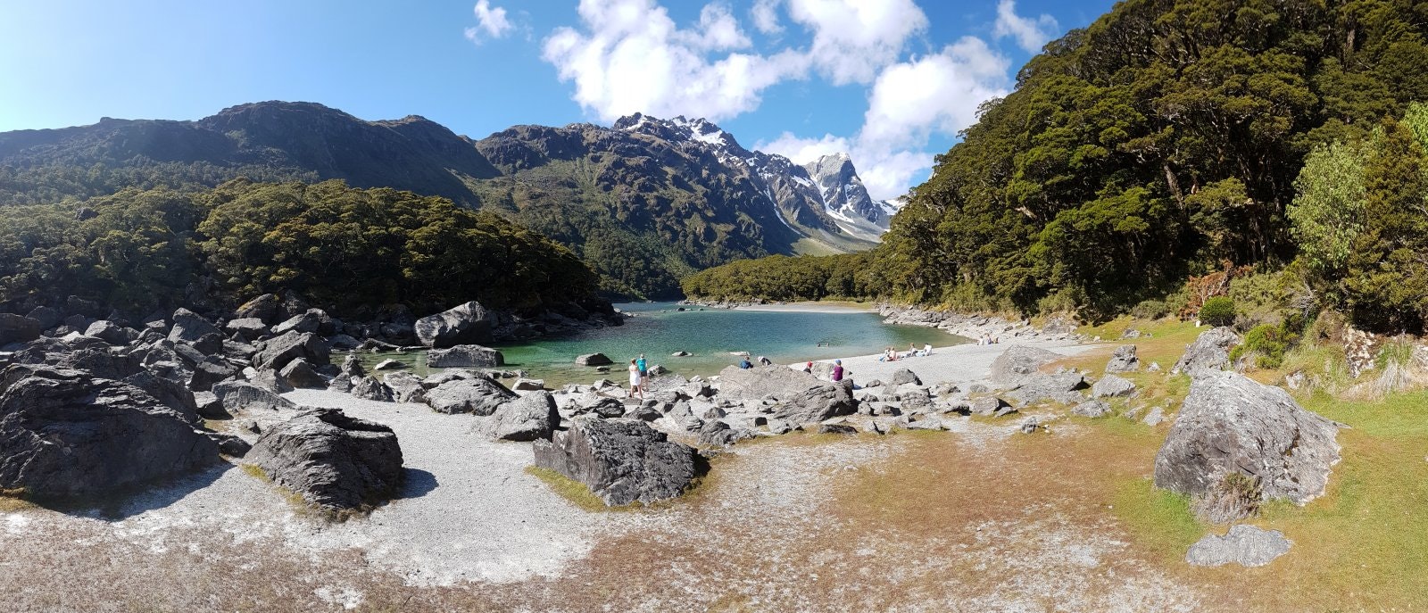 a panoramic shot of Lake MacKenzie on New Zealand's South Island on a sunny day. There are a few people near the water's edge standing and lying on a small stony beach. The blue water is surrounded by rocky, snow-capped mountains and lush greenery.