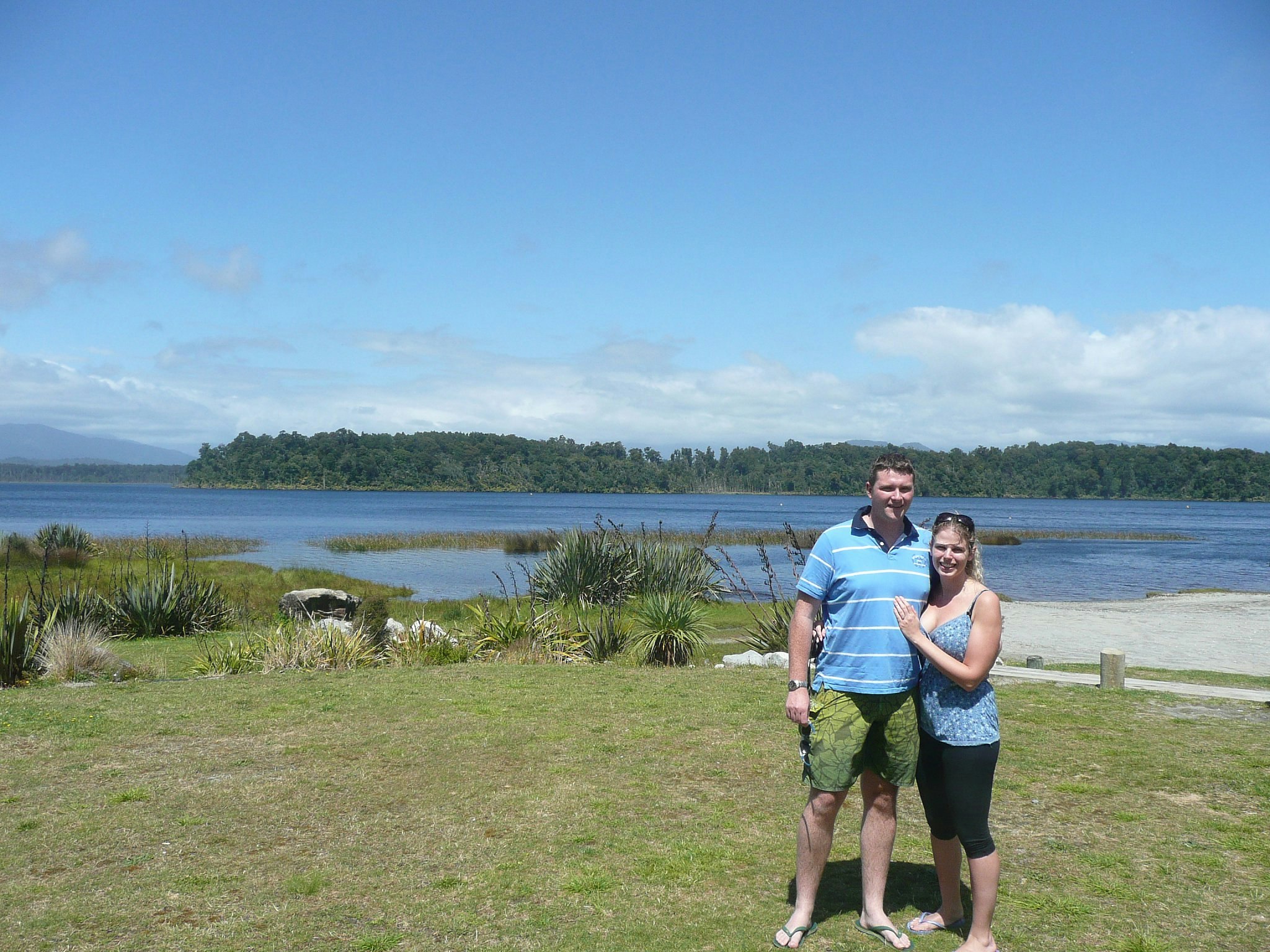 Lonely Planet staffer Becky and her partner stand in front of Lake Mahinapua, New Zealand on a sunny day; the far shore of the lake is tree-lined and there are mountains beyond.
