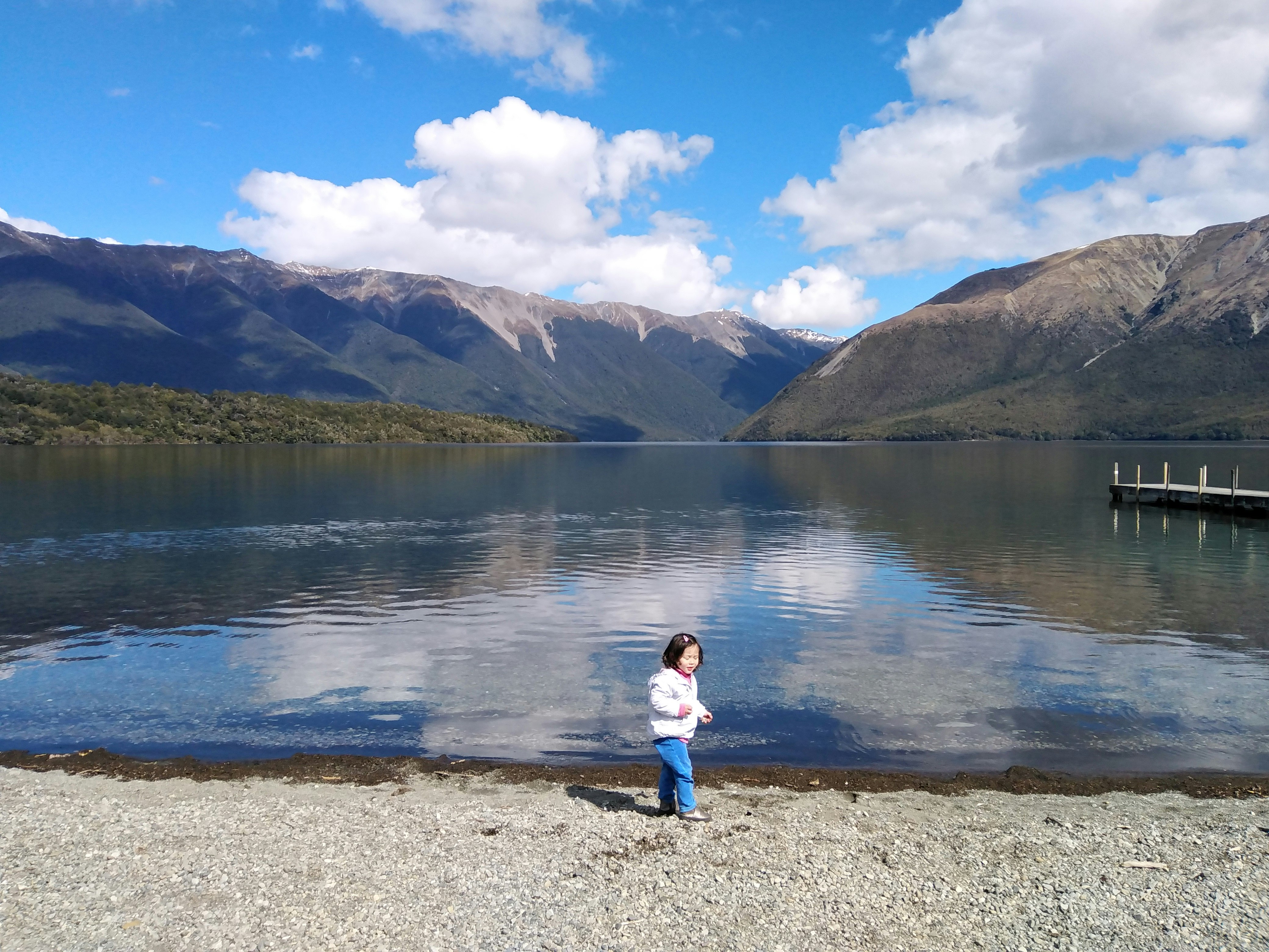A small child is standing at the bank of Lake Rotoiti, New Zealand. A group of mountains is visible in the background, as is a small pier to the right of the shot.