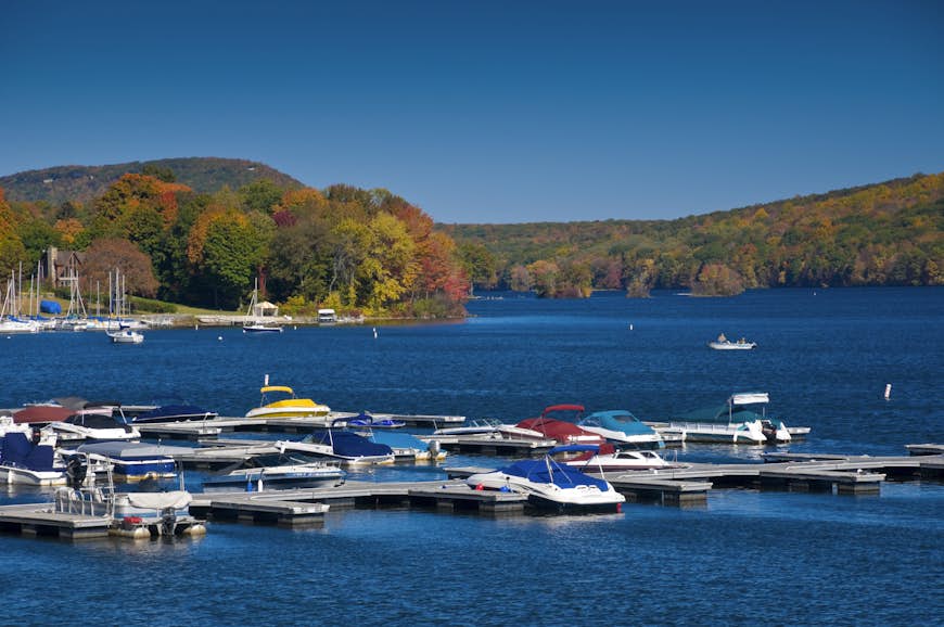 Boats are moored at a marina in a brilliant blue lake in autumn, with fall colors on the lake shores; New England fall foliage road trip