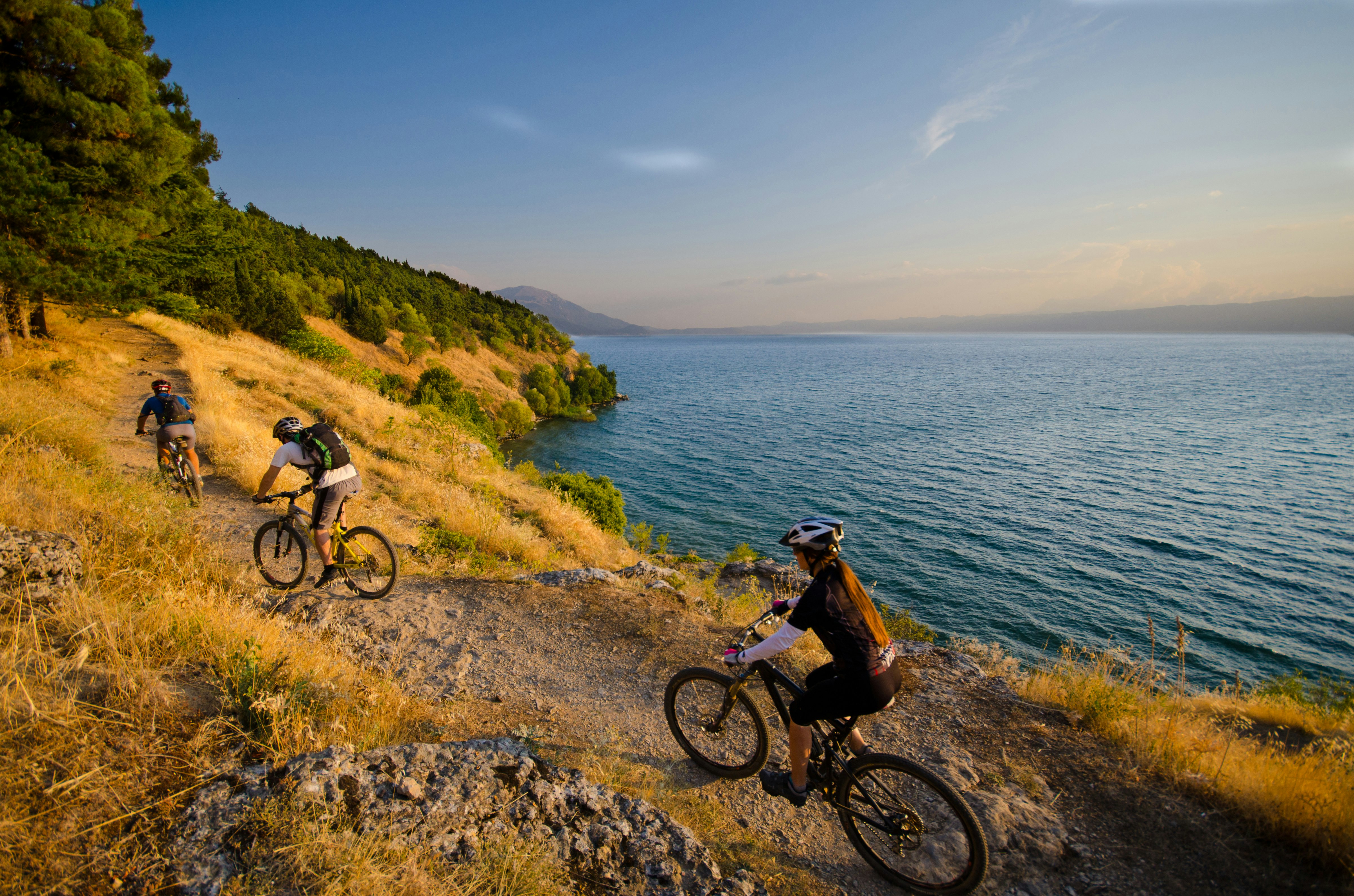 A trio of bike riders ascend uphill on a graveled path next to Lake Ohrid; North Macedonia Best in Travel