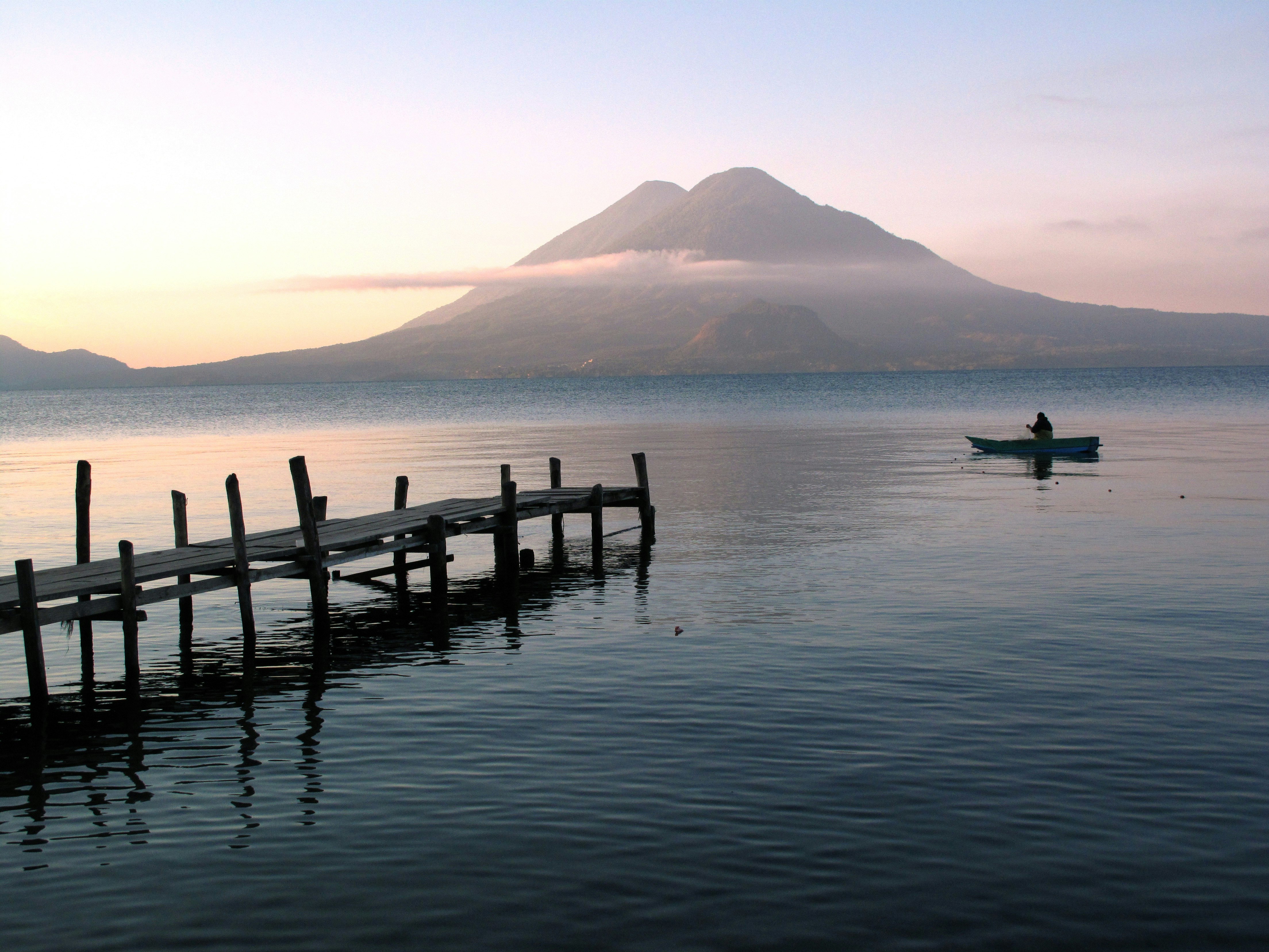 A rickety wooden harbour splinters out into the calm blue waters of Lake Atitlán. It's sunset, and a small, solitary boat glides through the water. In the background, on the opposite bank of the lake, a volcano is visible, silhouetted against the sunset.