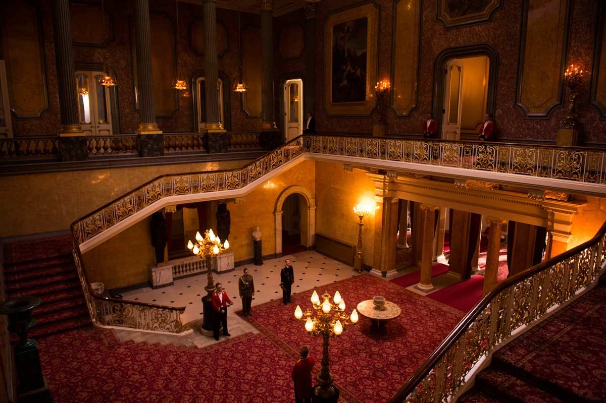 A still from The Crown, showing the ornate red-carpeted stairway at Lancaster House leading down to a hallway where staff are standing to attention.