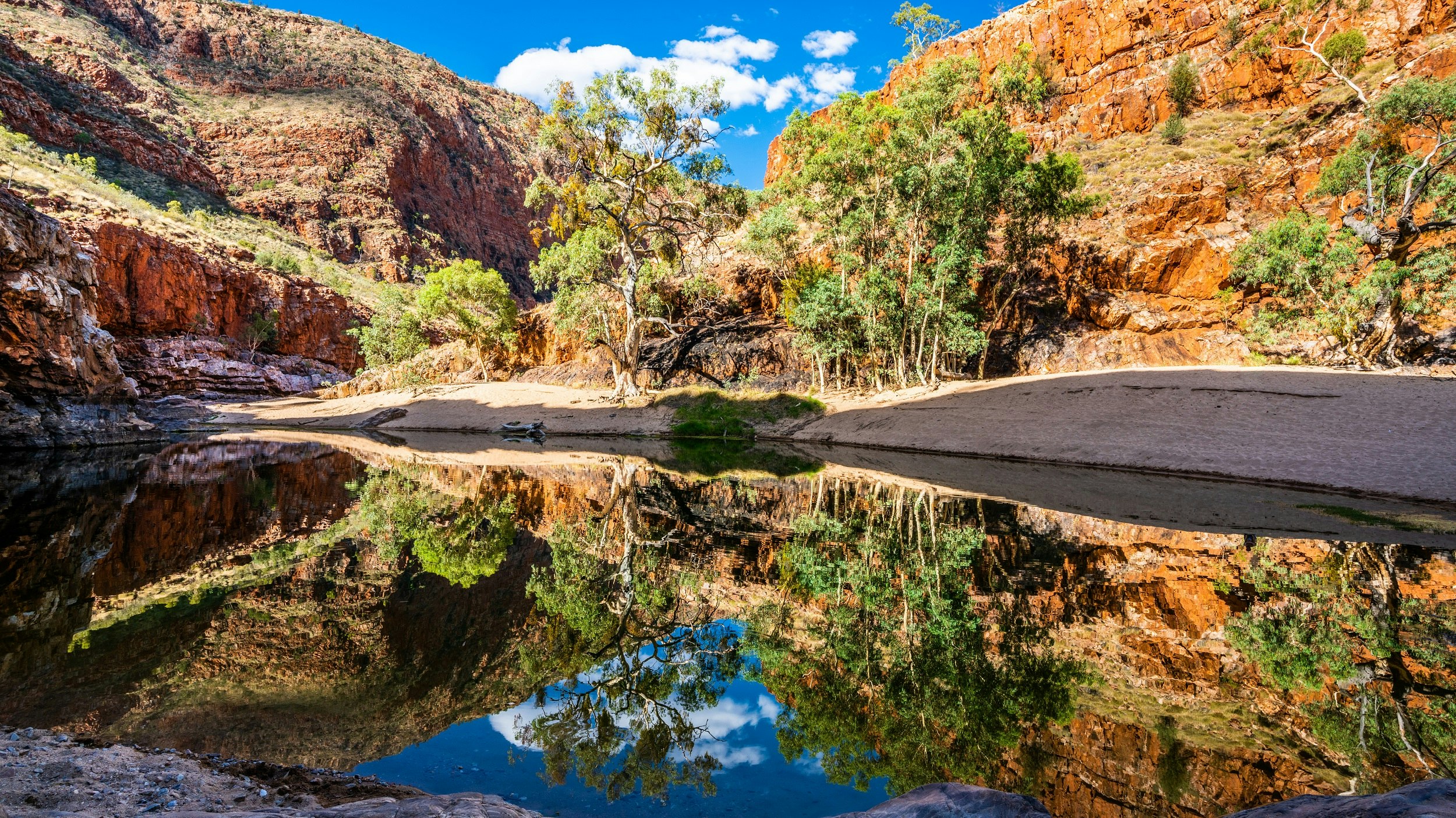 Ormiston gorge water hole in the West MacDonnell Ranges, Northern Territory; rec cliffs fringed with green grasses loom over a few trees and a waterhole, which reflects the scene and the blue sky above