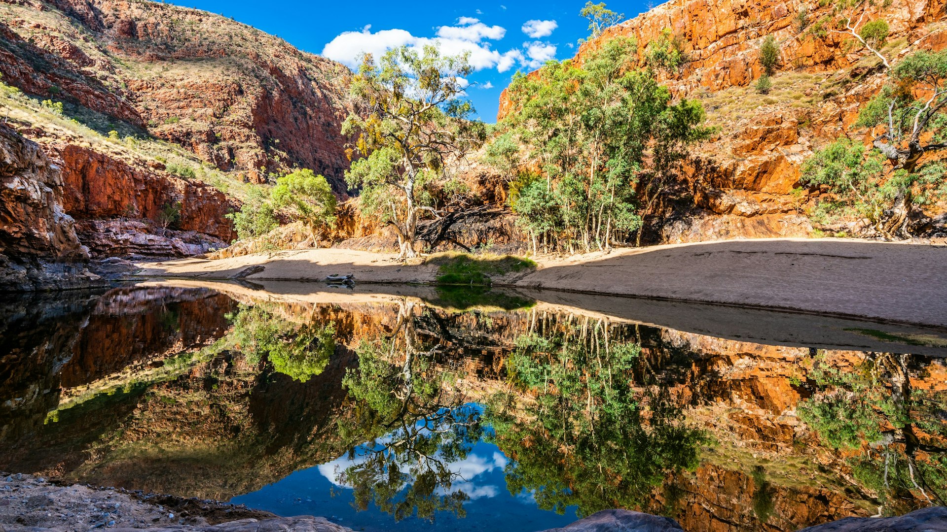 Ormiston gorge water hole in the West MacDonnell Ranges, Northern Territory; rec cliffs fringed with green grasses loom over a few trees and a waterhole, which reflects the scene and the blue sky above
