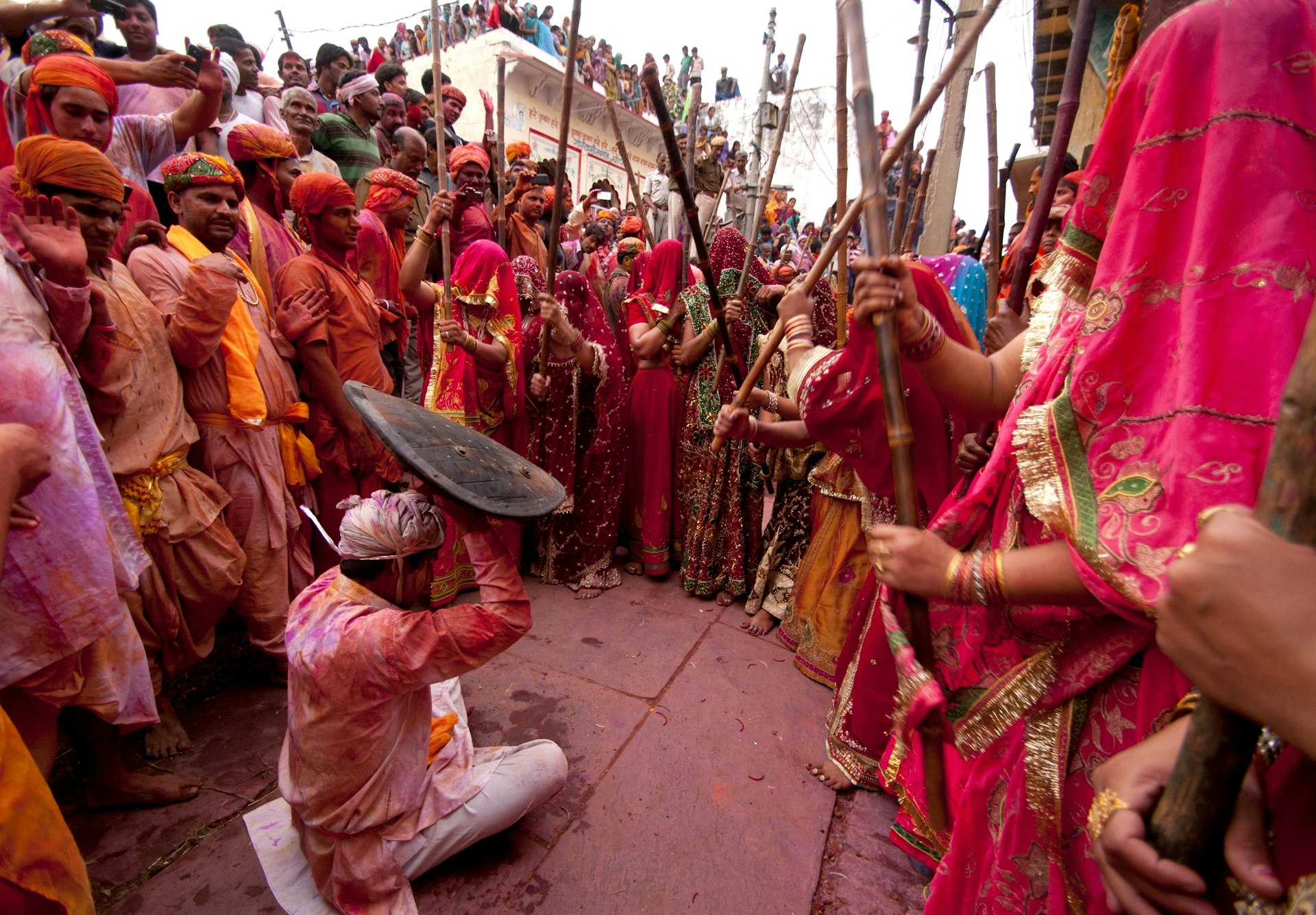 A group of women, all dressed in red saris, ceremoniously 'beat' a man, who cowers on the ground beneath a small shield, with large wooden sticks as part of Holi celebrations in India.