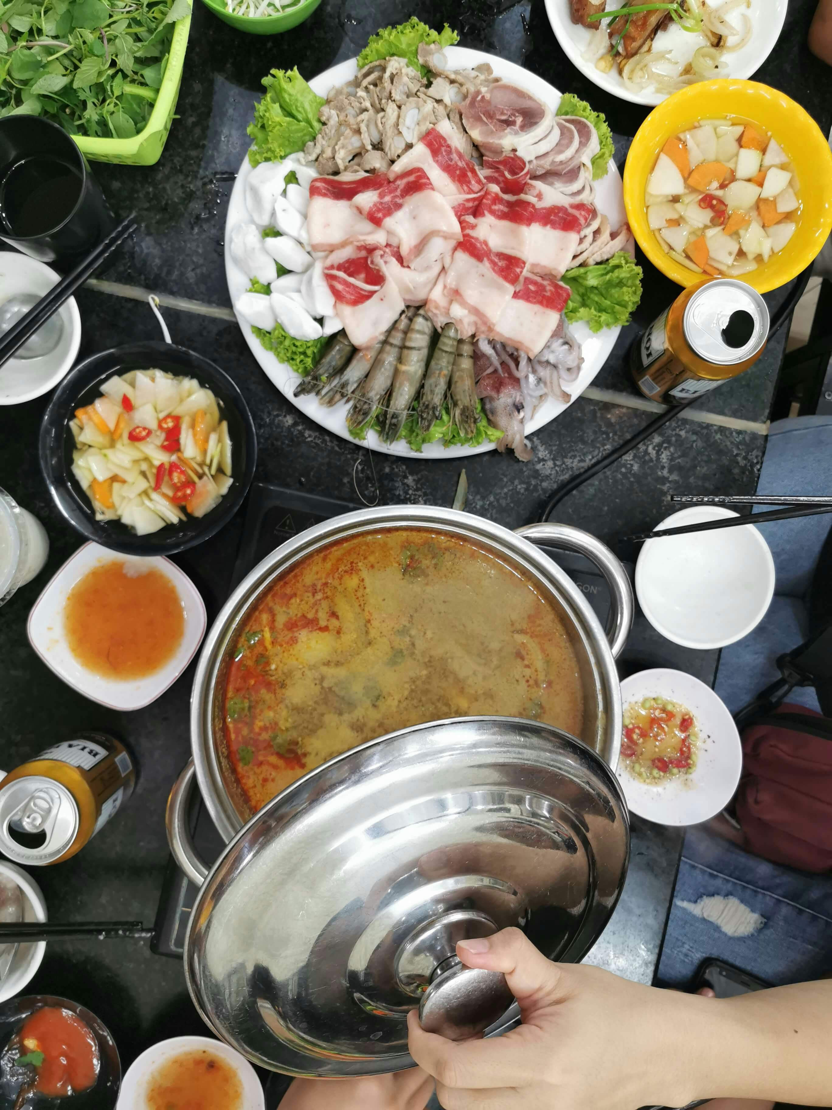 Someone is taking off a lid on a big pot of broth. Other dishes filled with vegetables and meat are on the table and two open cans of beer.