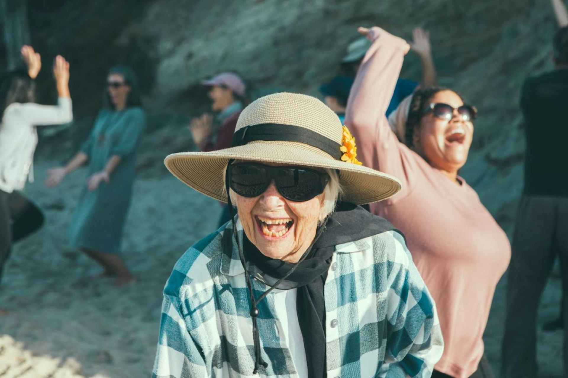 An elderly lady, wearing a hat and sunglasses, laughs in the direction of the camera, while participating in a laughter yoga class in Laguna Beach, California.