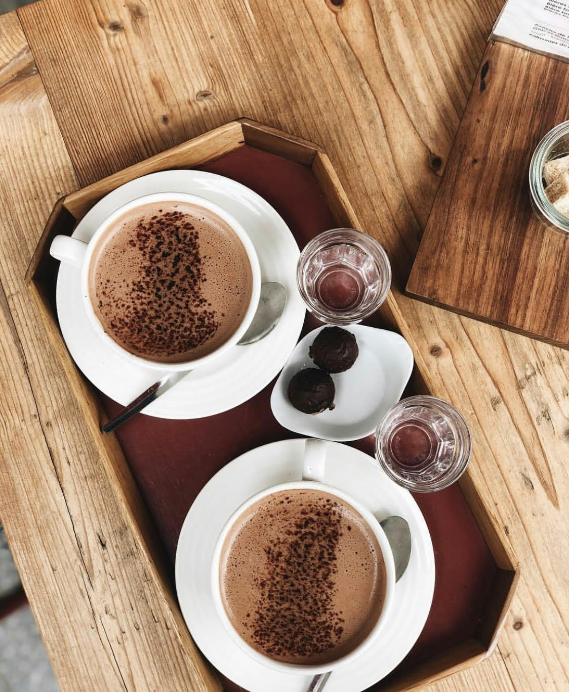 Plain white mugs of hot chocolate sprinkled with spices and more chocolate on wooden trays arranged on warm wood tables with knots and wood grain visible.