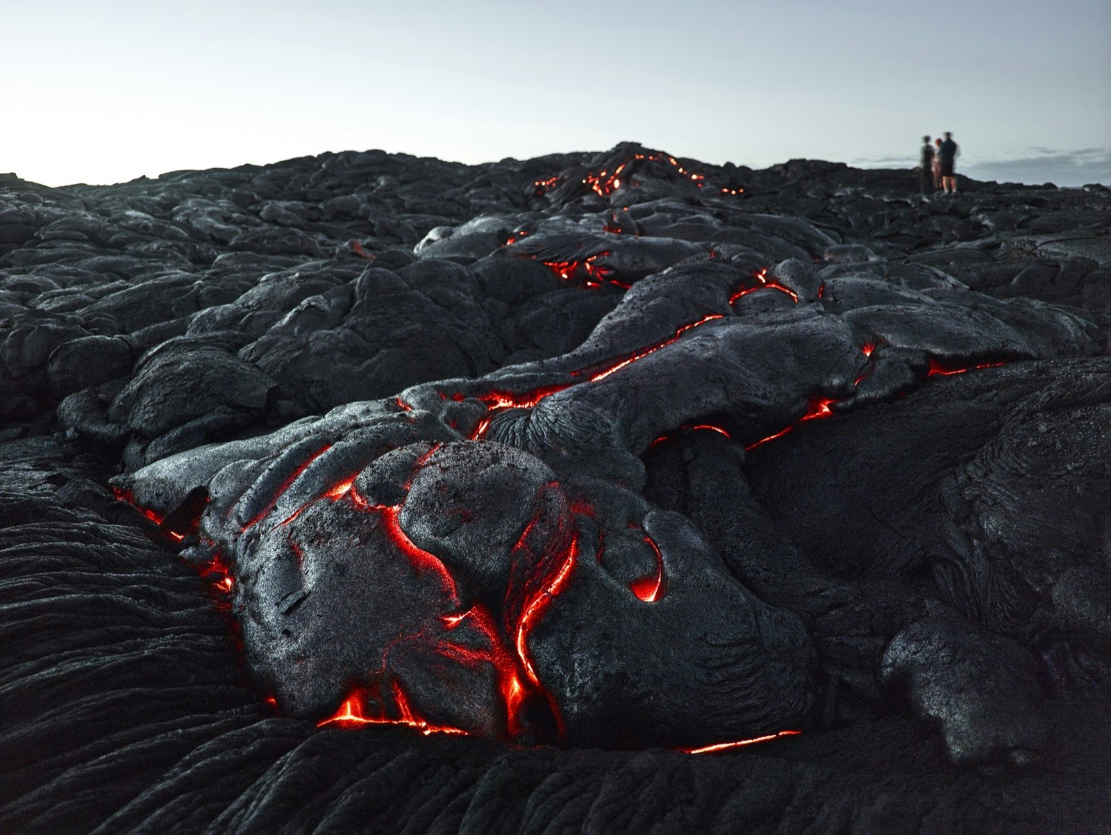 People stand in the background as hot red lava flows through the cracks of black hardened lava