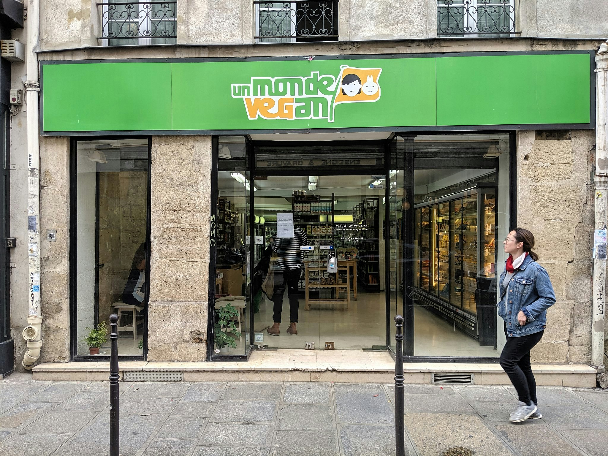 A woman walks past in a shop housed in a historic stone building; a green sign says Un Monde Vegan. 