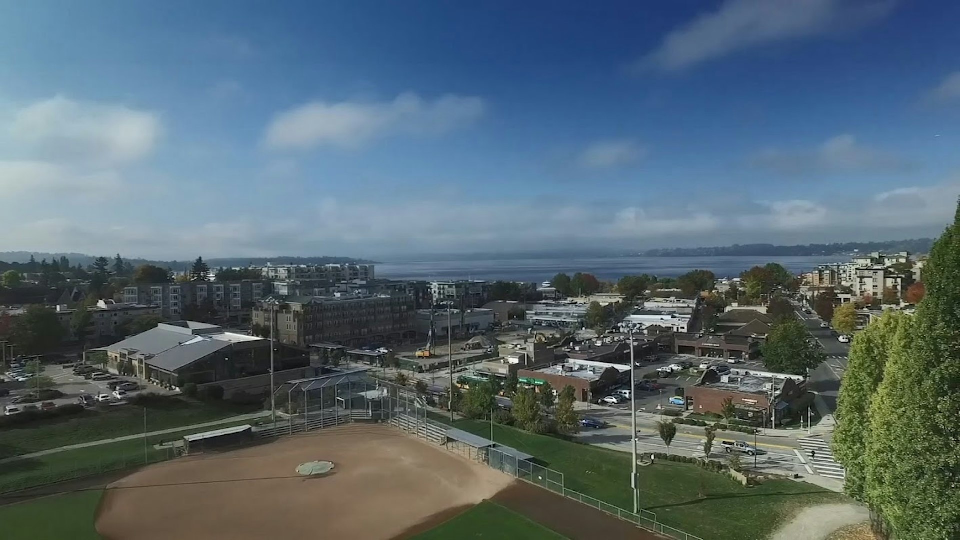 This shot overlooks the clean brown baseball diamond at Lee Johnson Field in Kirkland from a high enough angle to afford a view of the surrounding low brick buildings and beyond them, the blue water of Lake Washington's Moss Bay