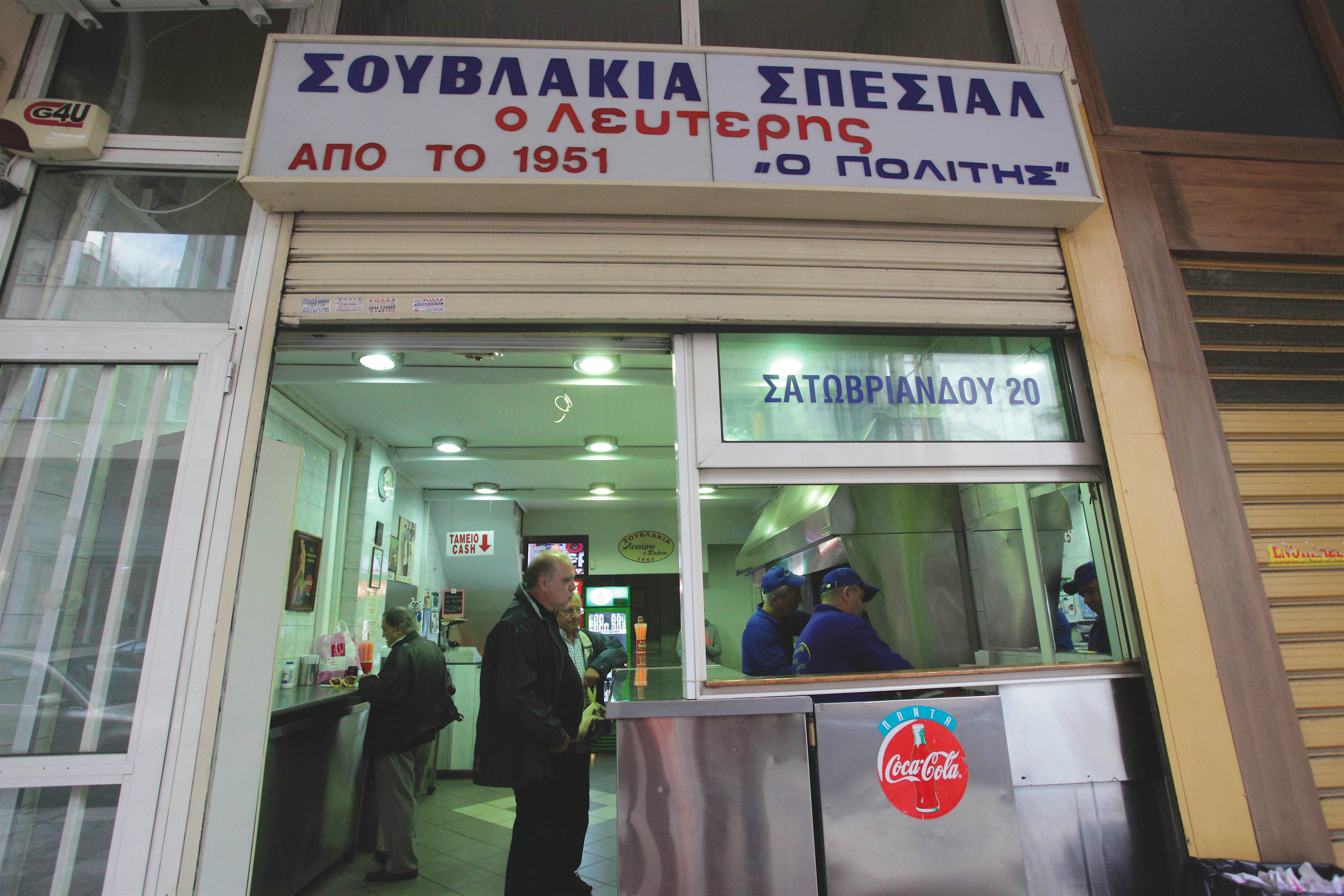 Lefteris-athenian-restaurants.jpg An older gentleman in black pants and a black coat waits at the counter of the Lefteris suvlaki stand in Athens, Greece while two employees in blue shirts prepare food in a stainless steel kitchenette