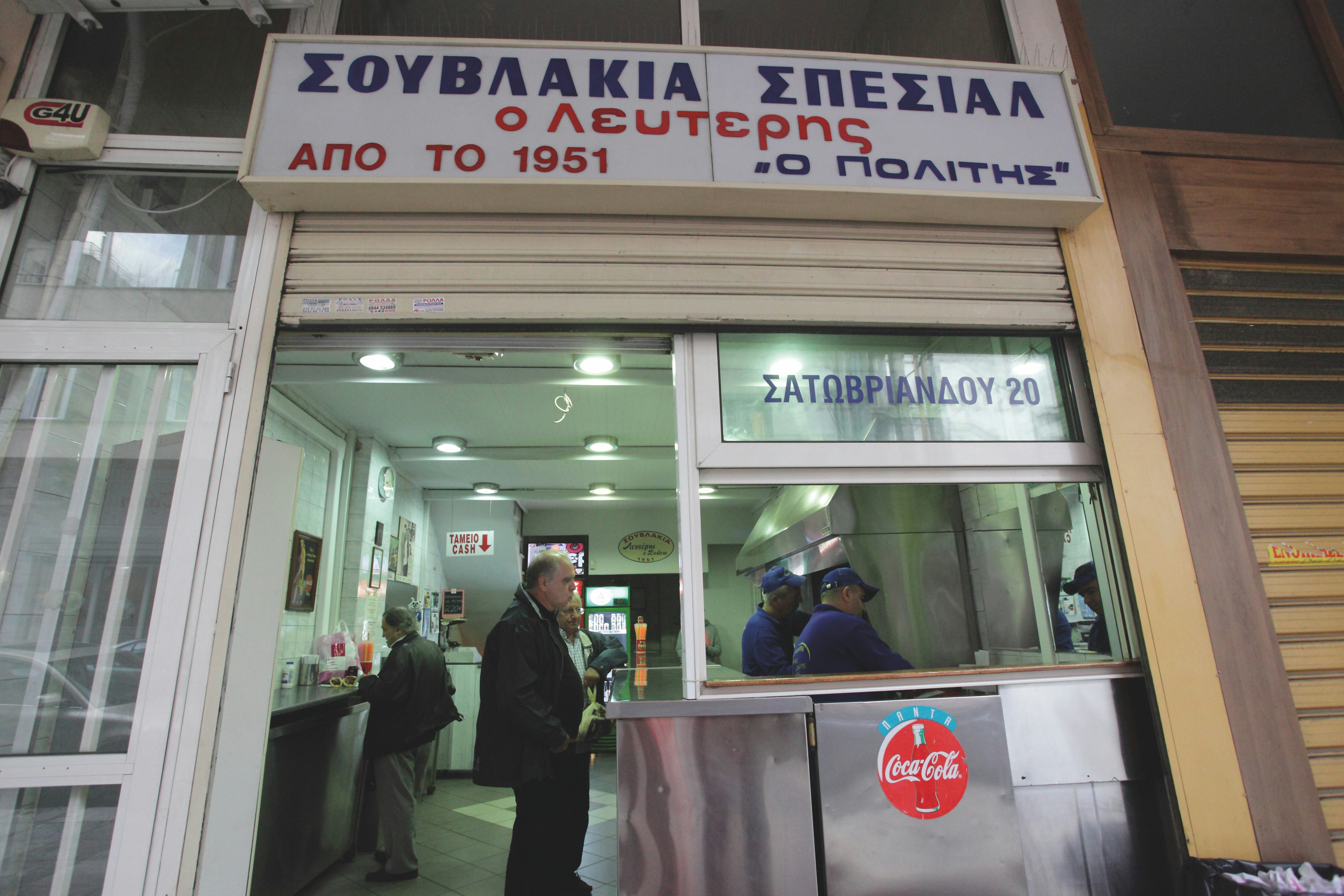 An older gentleman in black pants and a black coat waits at the counter of the Lefteris suvlaki stand in Athens, Greece while two employees in blue shirts prepare food in a stainless steel kitchenette 