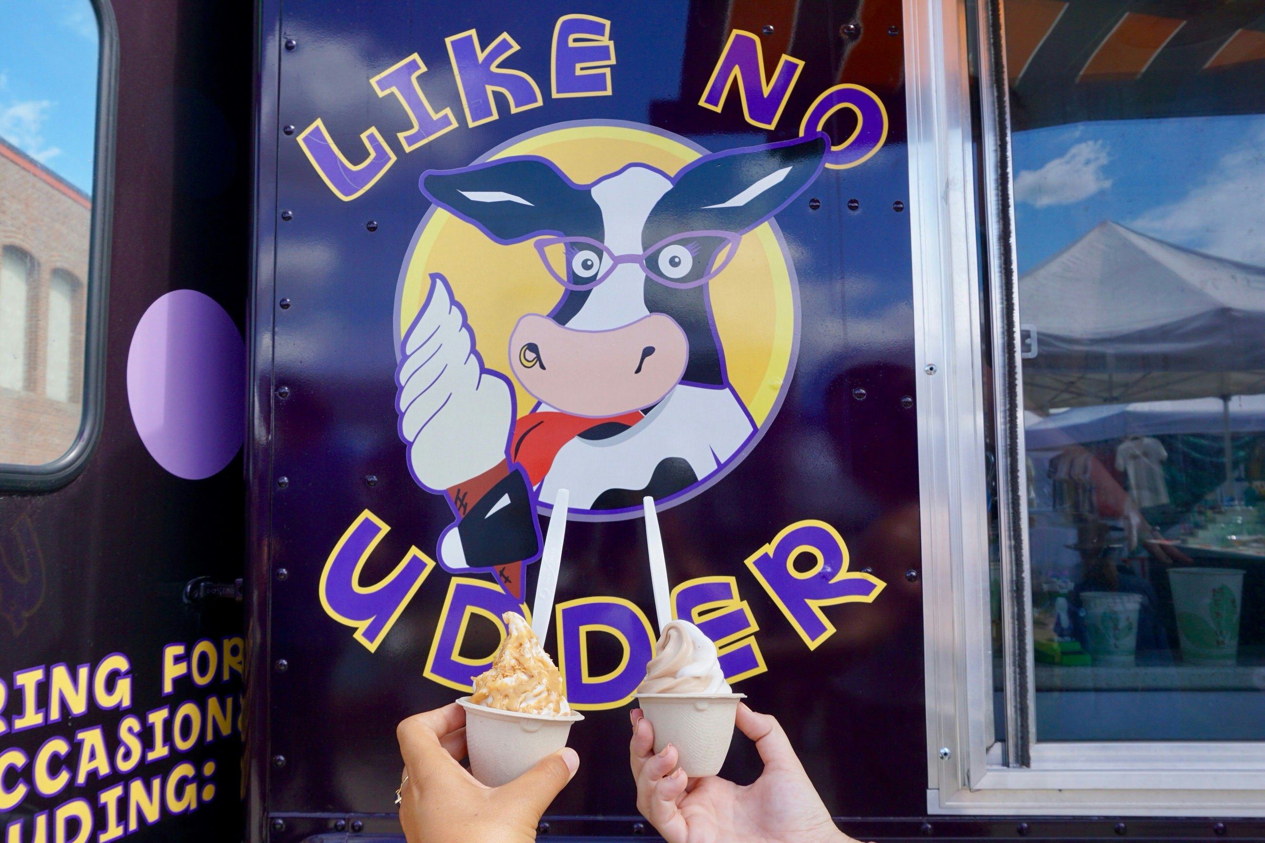 Two people are holding up servings of vegan soft-serve ice cream in the frame - only their hands can be seen in front of the 'Like No Udder' sign on the side of an ice-cream truck. The sign is a cartoon cow wearing glasses and licking an ice-cream cone.