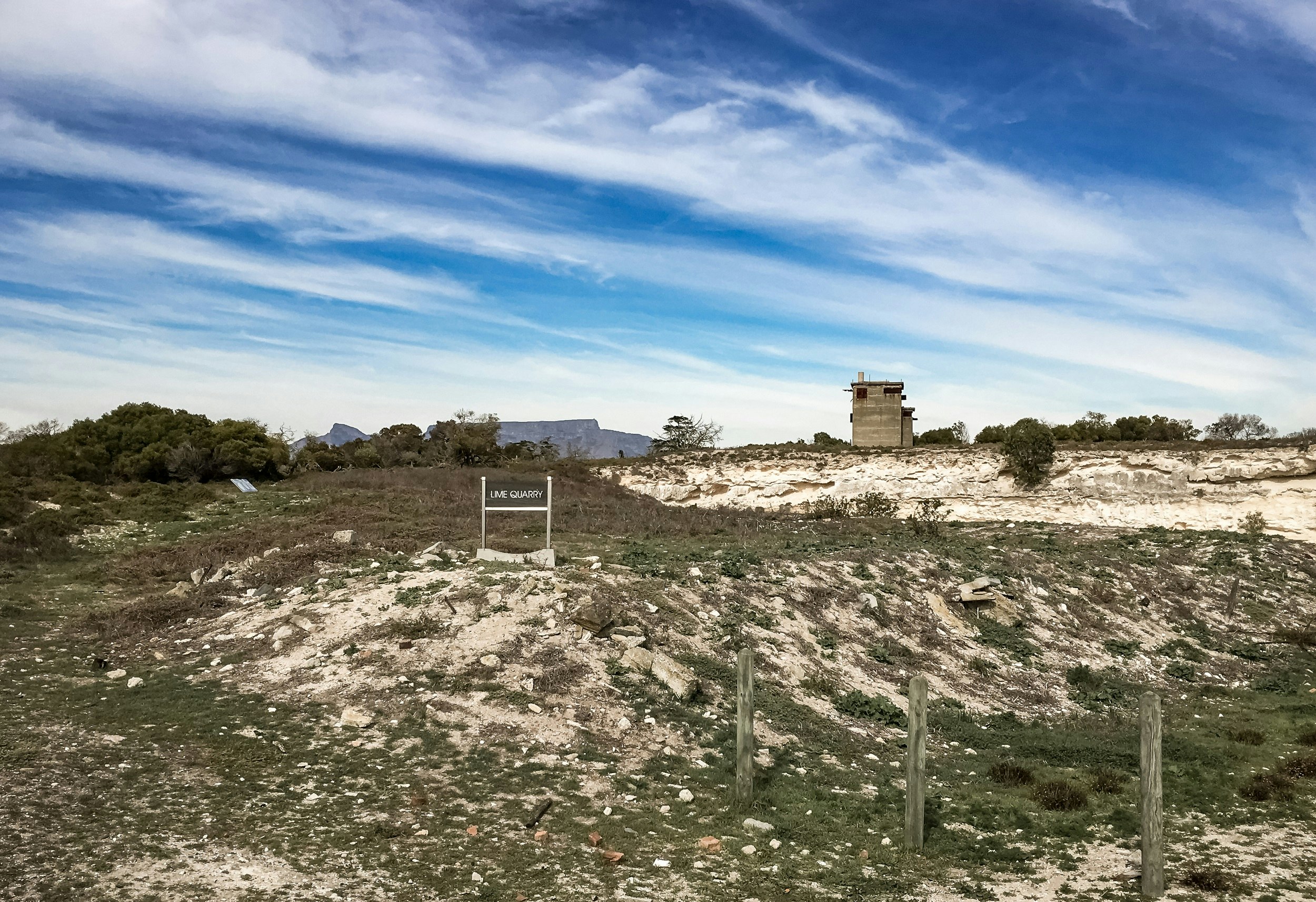 Bright limestone walls of a rock quarry; in the foreground is shrubby vegetation on the base of the quarry, while atop the walls are trees and a concrete guard tower.