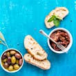Mediterranean snacks set. Olives, oil, sun-dried tomatoes, herbs and sliced ciabatta bread on over blue painted background, top view