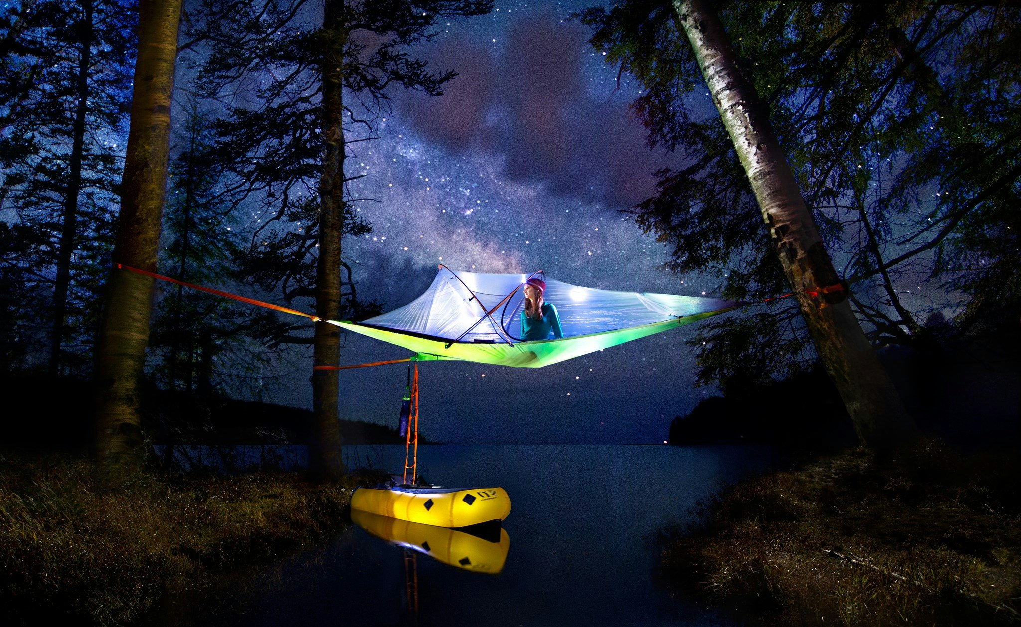 A woman sits perched in a Tentsile tree tent over a watery inlet. The Tentsile is illuminated against the night sky, and you can see she's moored a yellow raft to the tent, which is fastened to three nearby trees.