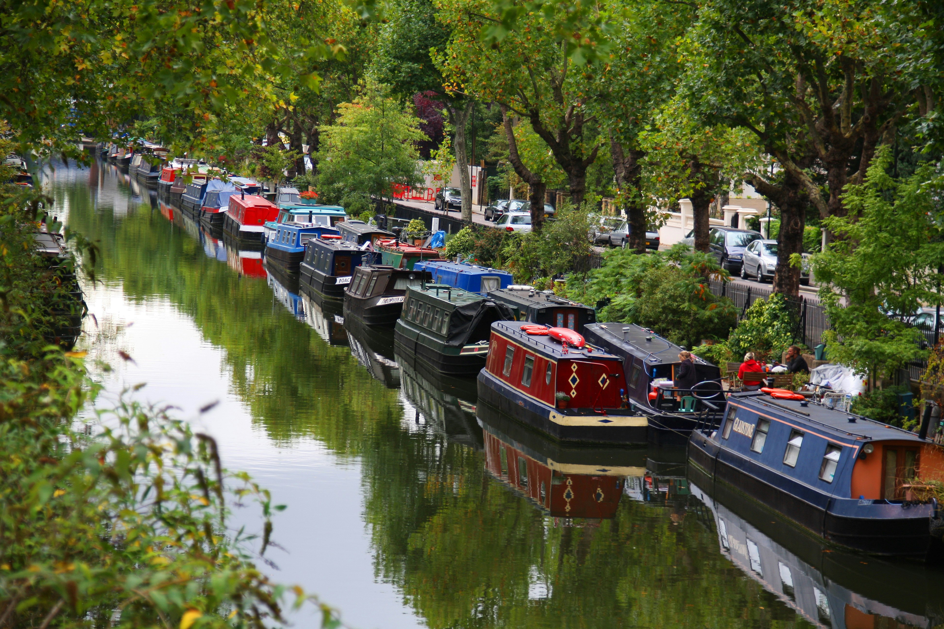 Two rows of house boats line the edge of a canal wall. Trees flank the paved walkway and a group of people sit on a wooden bench; Heathrow Layover 
