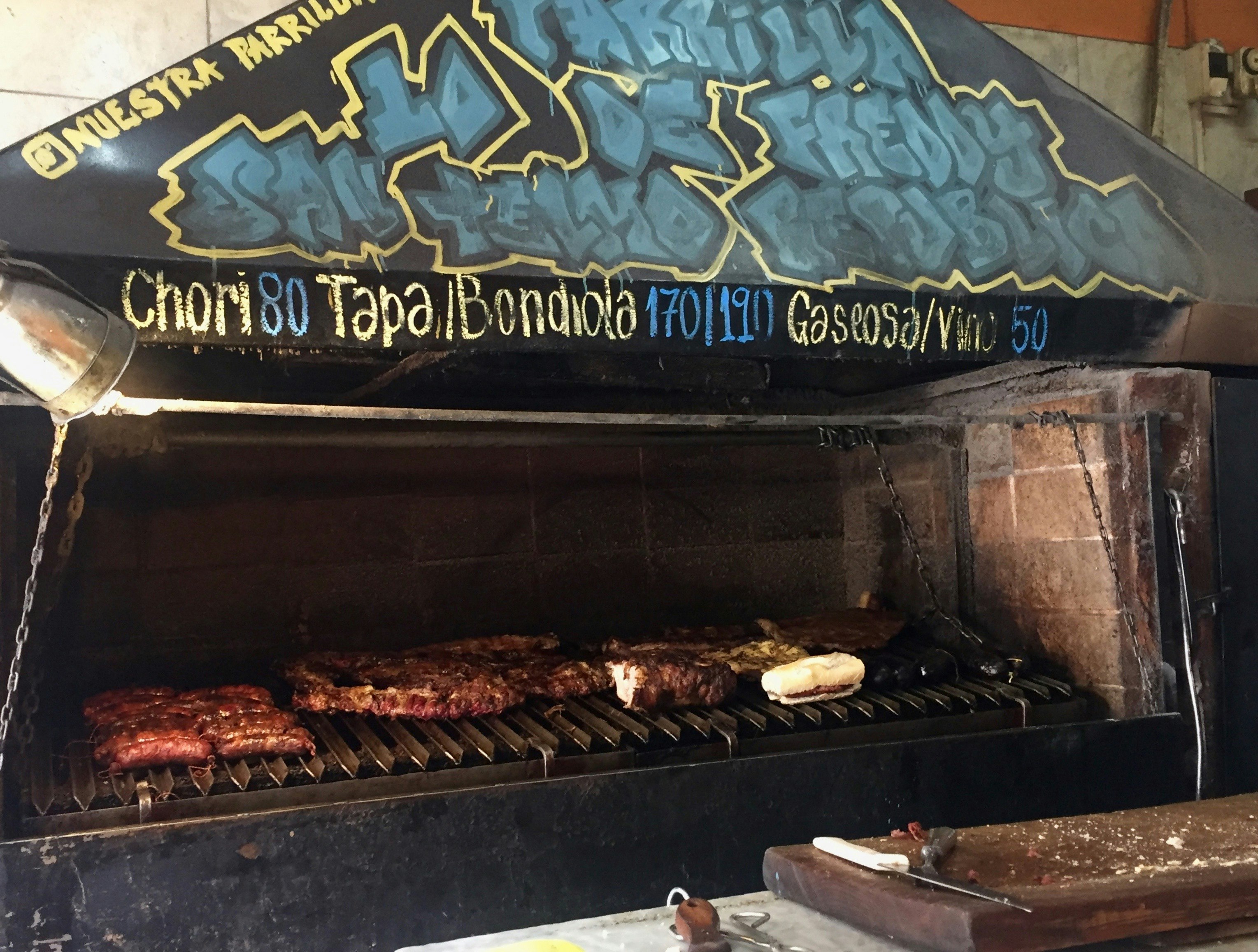 Steaks and sausages cooking on the very old and well-used grill at Lo de Freddy. The grill hood has the stand's details in blue graffiti.
