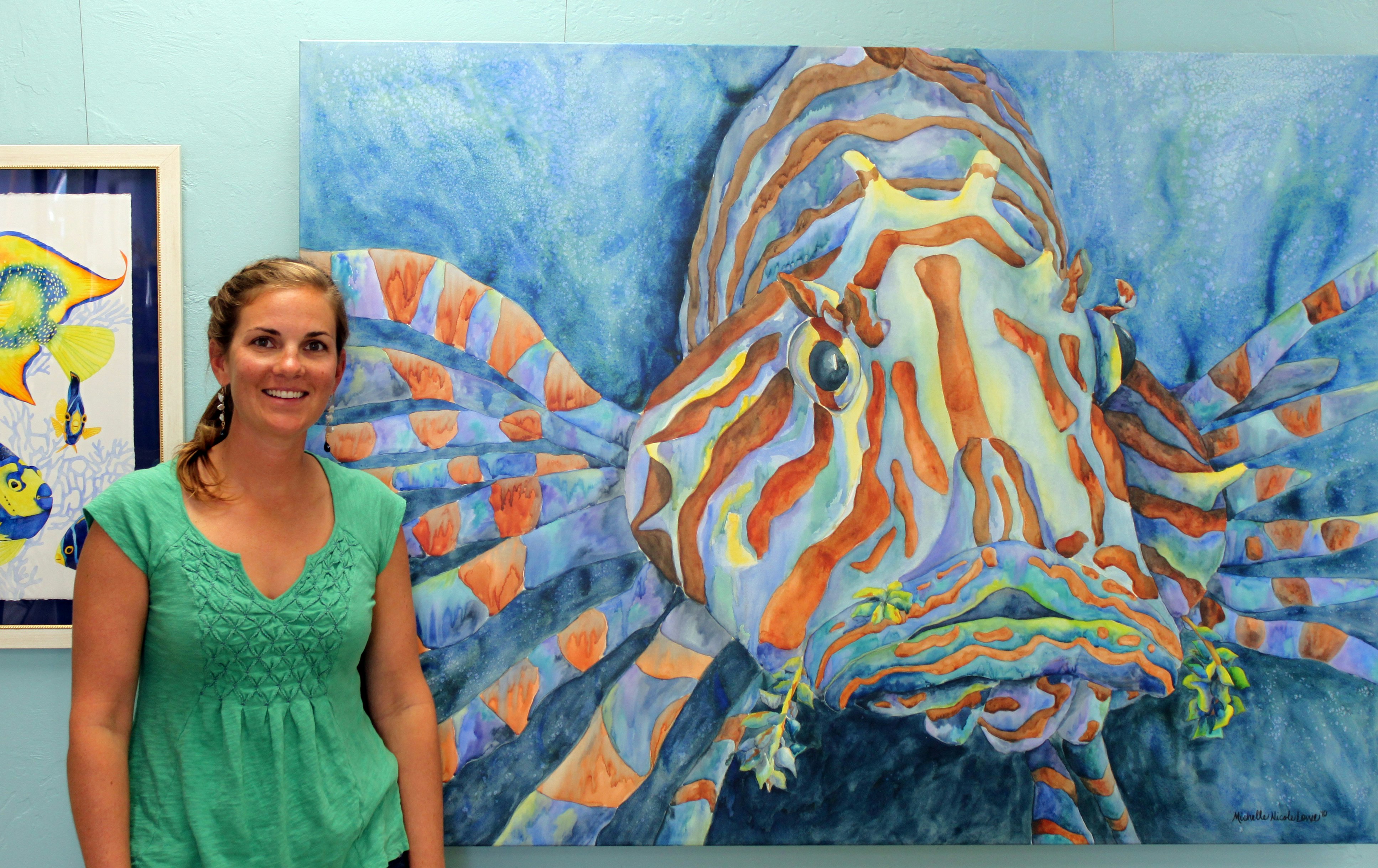 A blonde woman in a green blouse smiles as she stands next to her painting, a large watercolor canvas depicting a large colorful lionfish