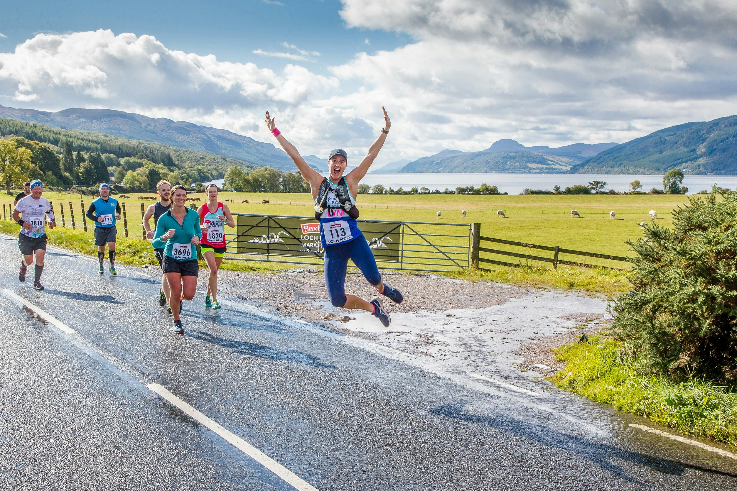 Half a dozen competitors run along a wet section of road along a farmer's field dotted with sheep; one runner is leaping in the air, with arms raised and heels clicking together. In the background are rolling green hills and Loch Ness.