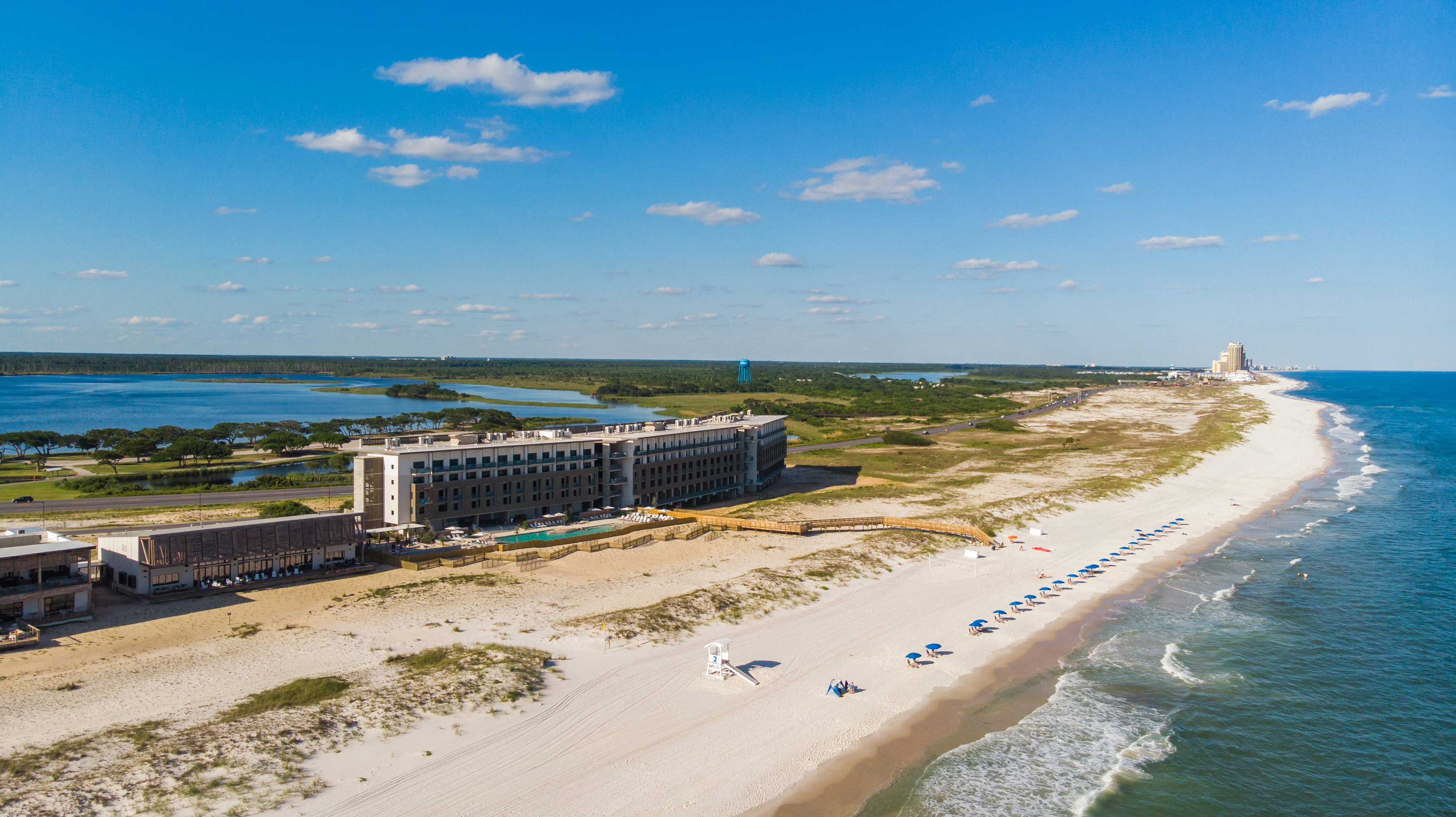 A strip of near-white sand runs alongside a grassy coastline; there is a very large but understated modern hotel set back from the beach. Behind this a lake can be seen.