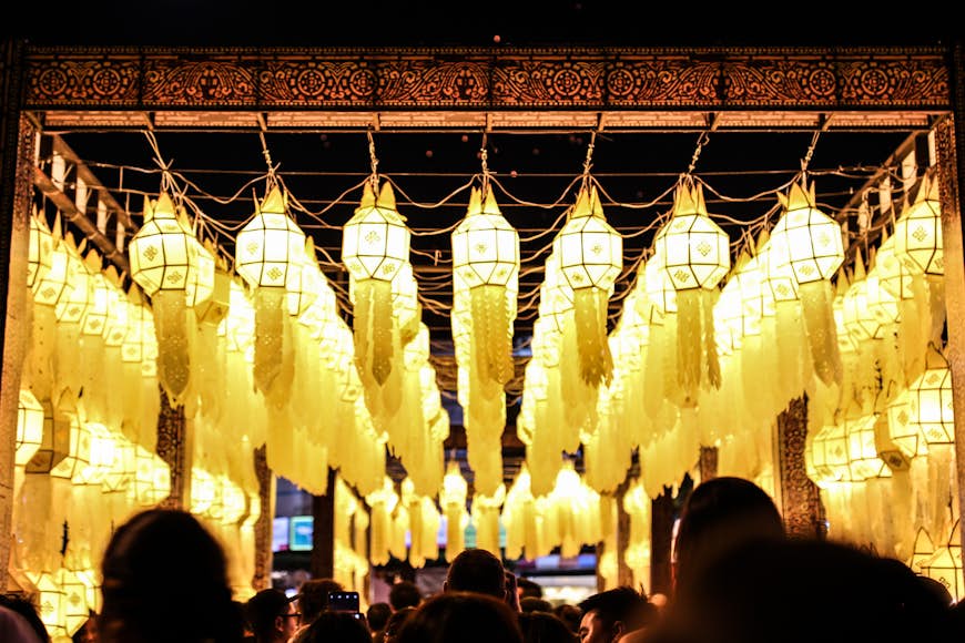 Illuminated yellow lanterns dangle on strings over a street in Chiang Mai. Beneath them, crowds of people watch on, with only their silhouettes visible.