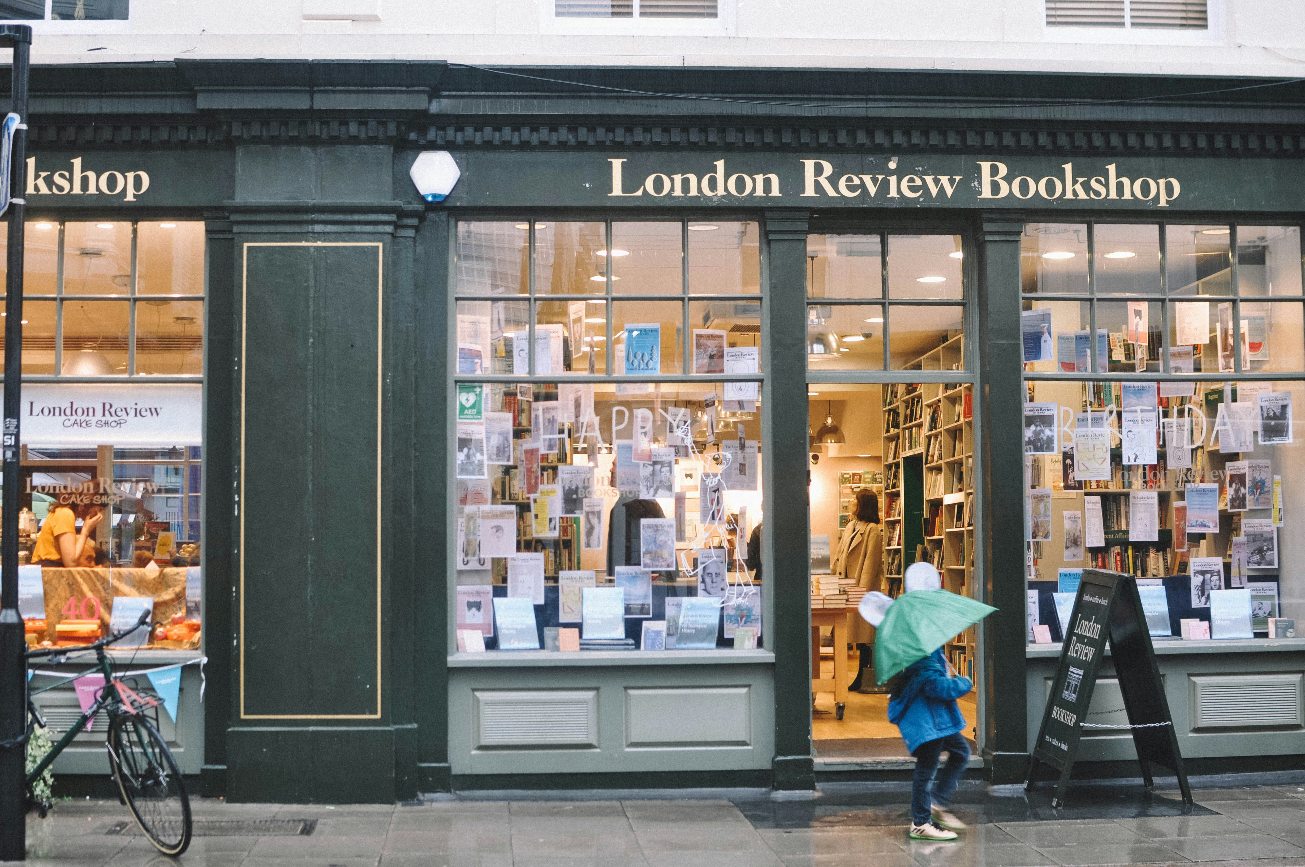 The outside of the London Review Bookshop. It's raining and a child walks past with a green umbrella. The inside of the store is lit up in a warm yellow and there are customers inside.