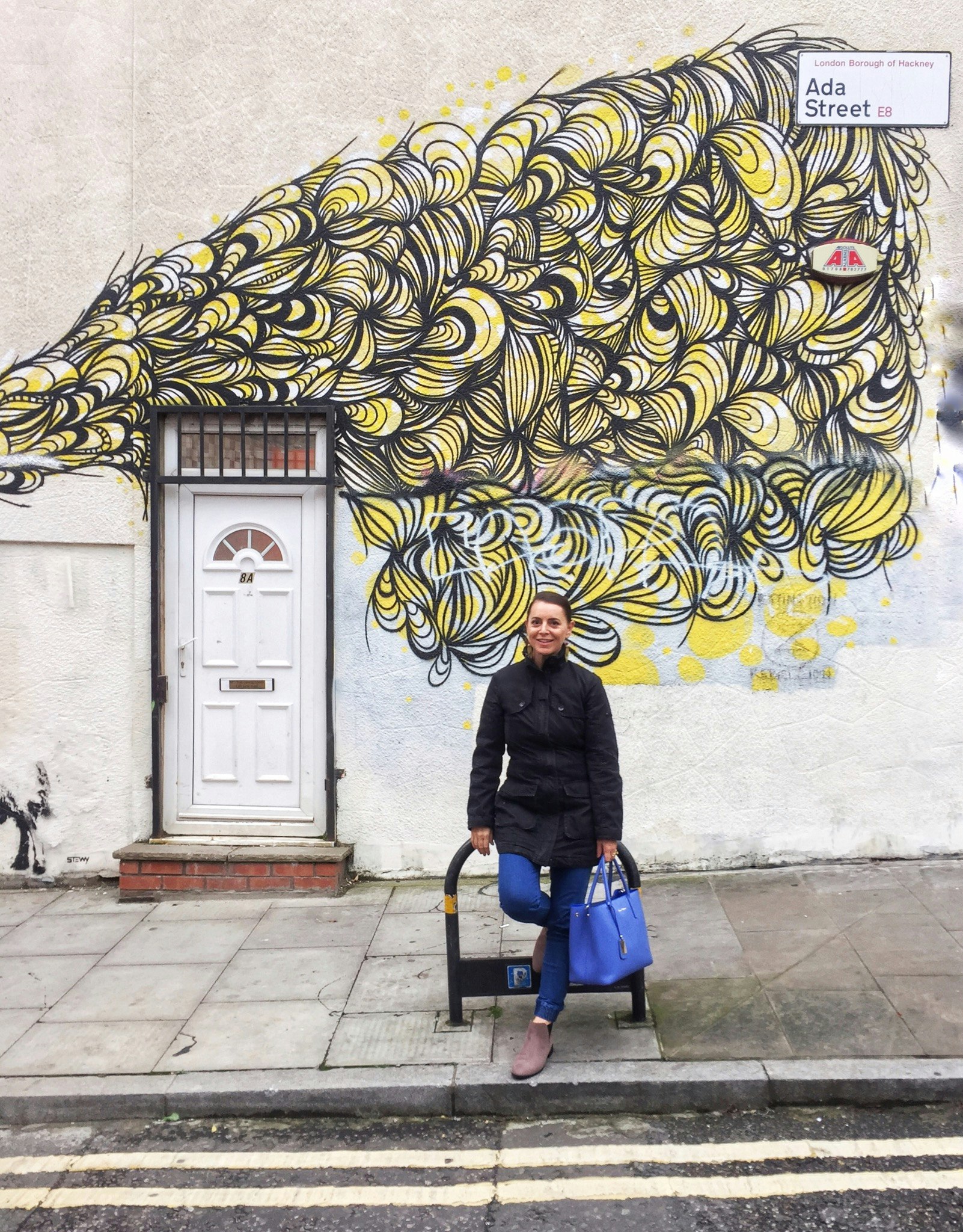 The writer, Nardia, poses in front of a yellow and black mural on Ada Street in east London.