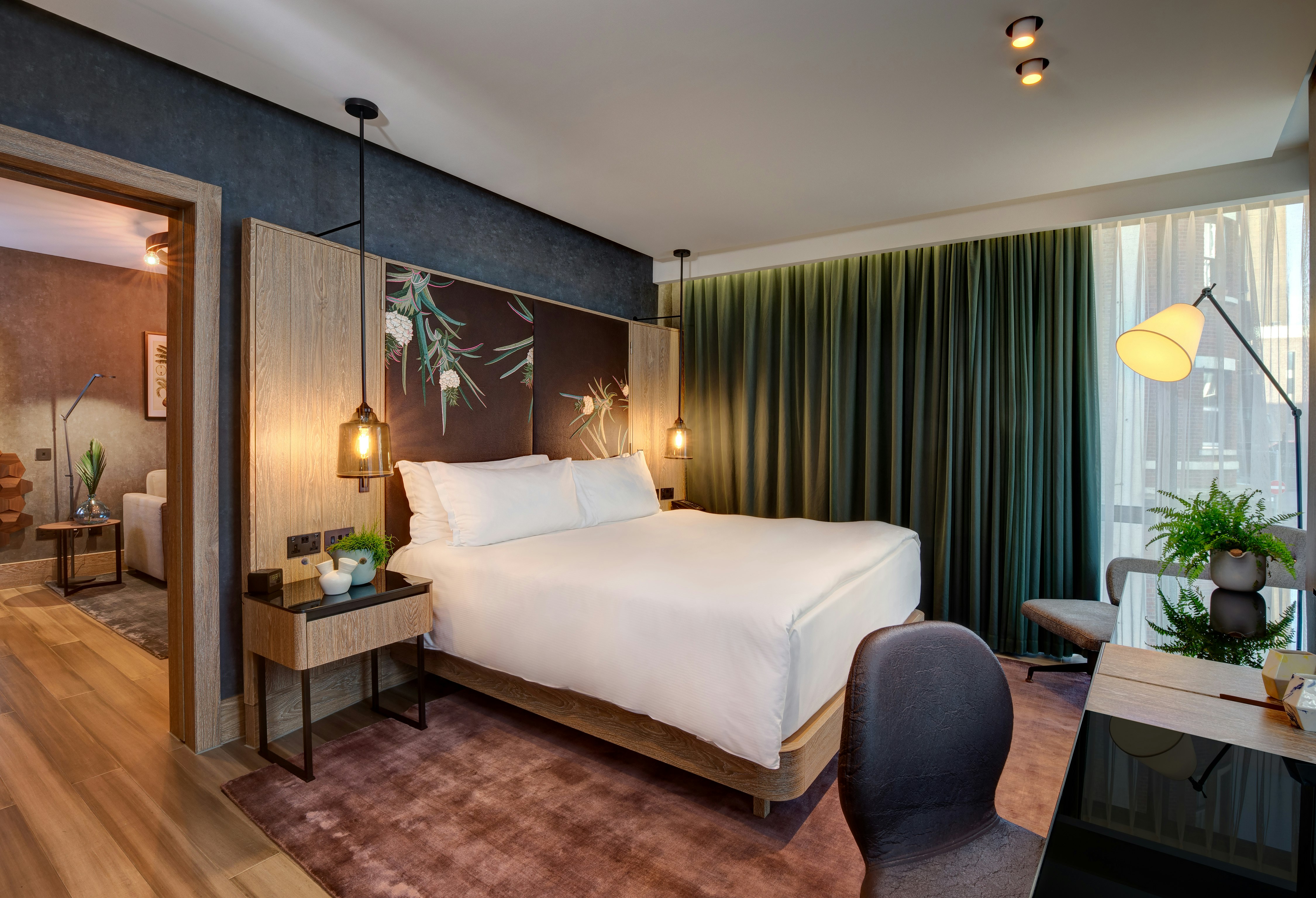 Exterior shot of a hotel room at the Hilton London Bankside. There's a bed covered in white covers and pillows, a wooden side table, a wooden chair and a wooden desk. There is a green curtain hanging over the large window. 
