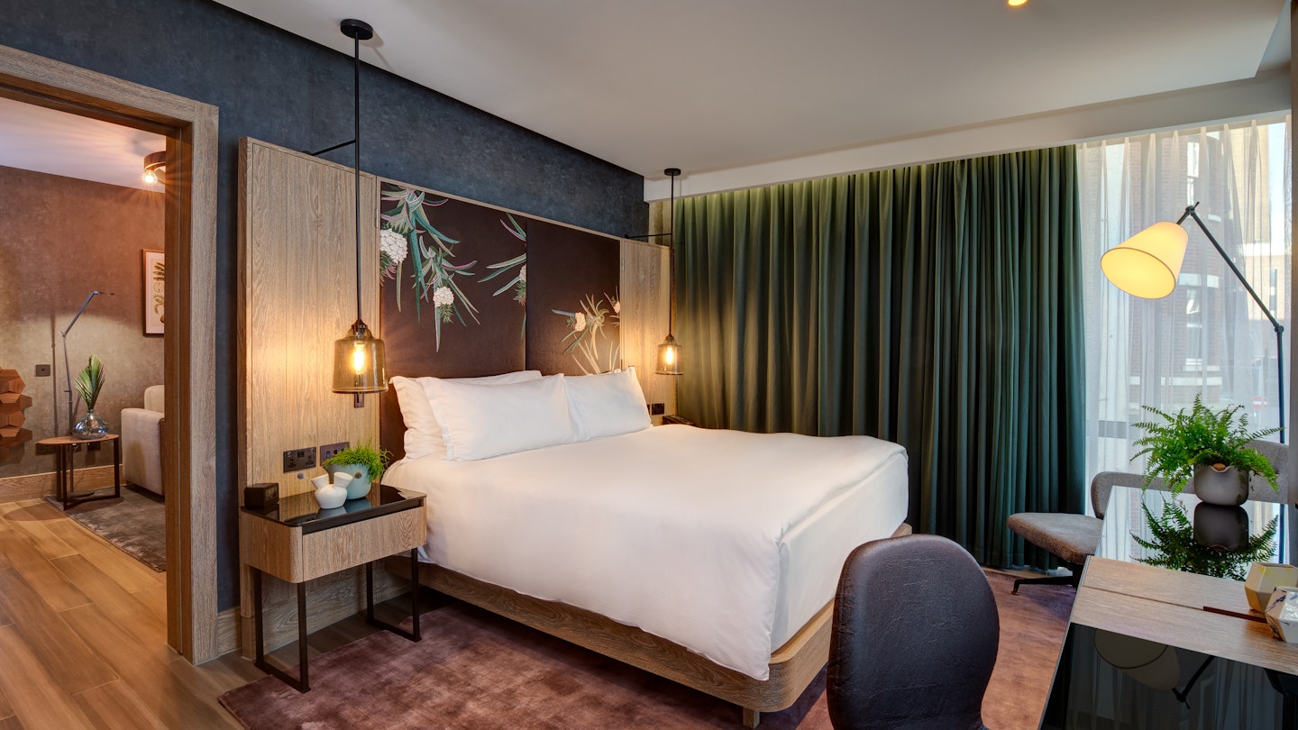 Exterior shot of a hotel room at the Hilton London Bankside. There's a bed covered in white covers and pillows, a wooden side table, a wooden chair and a wooden desk. There is a green curtain hanging over the large window. 
