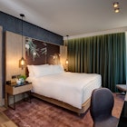 Exterior shot of a hotel room at the Hilton London Bankside. There's a bed covered in white covers and pillows, a wooden side table, a wooden chair and a wooden desk. There is a green curtain hanging over the large window.
