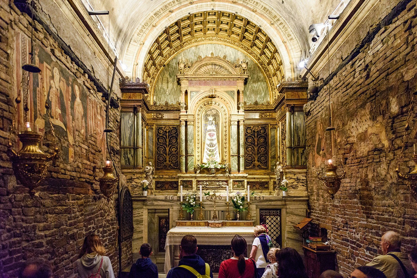 People stand in a narrow room with old brick walls, a vaulted ceiling, and a golden altar at the Basilica Santa Casa. Le Marche, Italy.