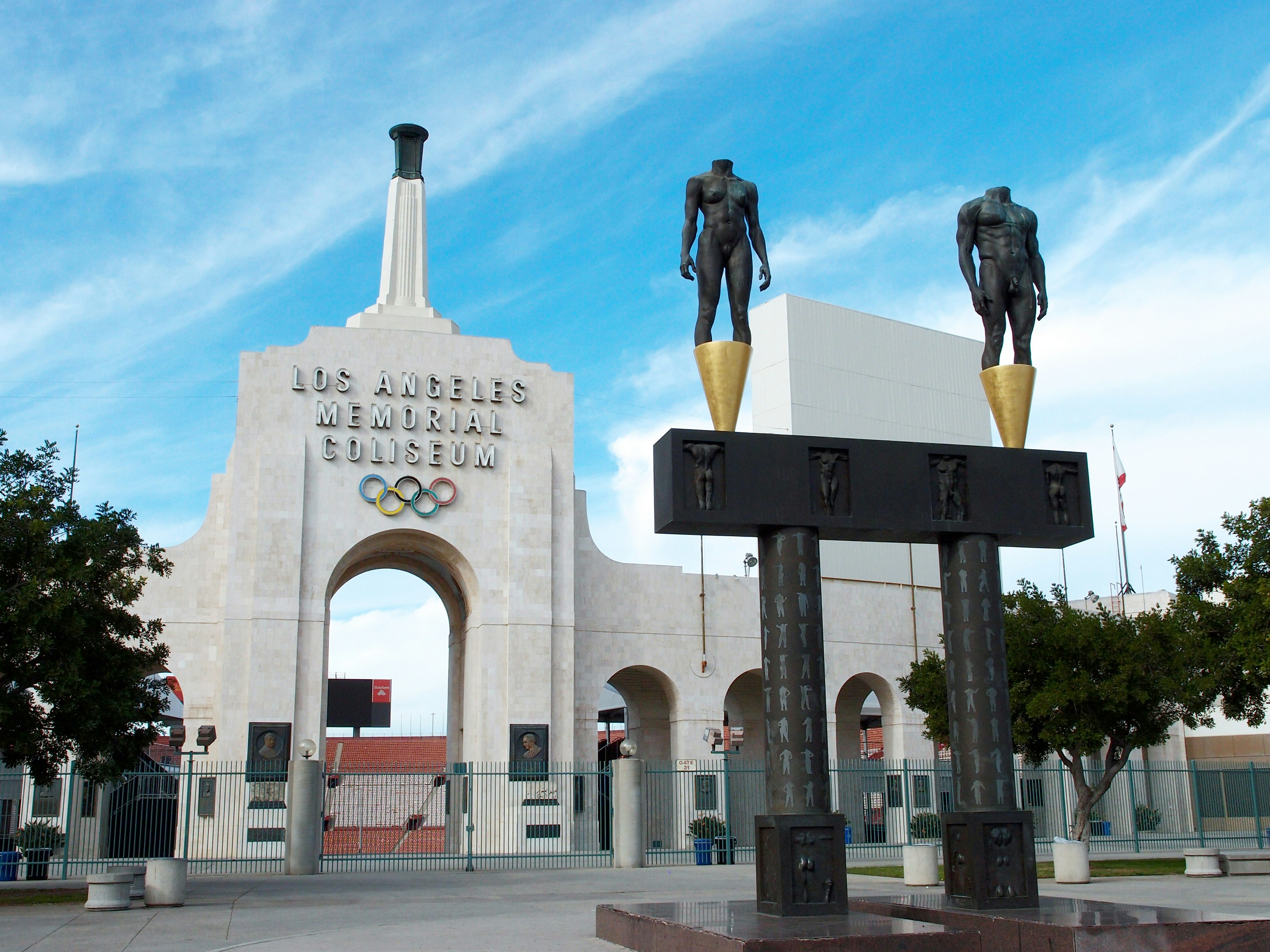Exterior shot of the Los Angeles Coliseum. There are a pair of headless statues standing on golden cones. 