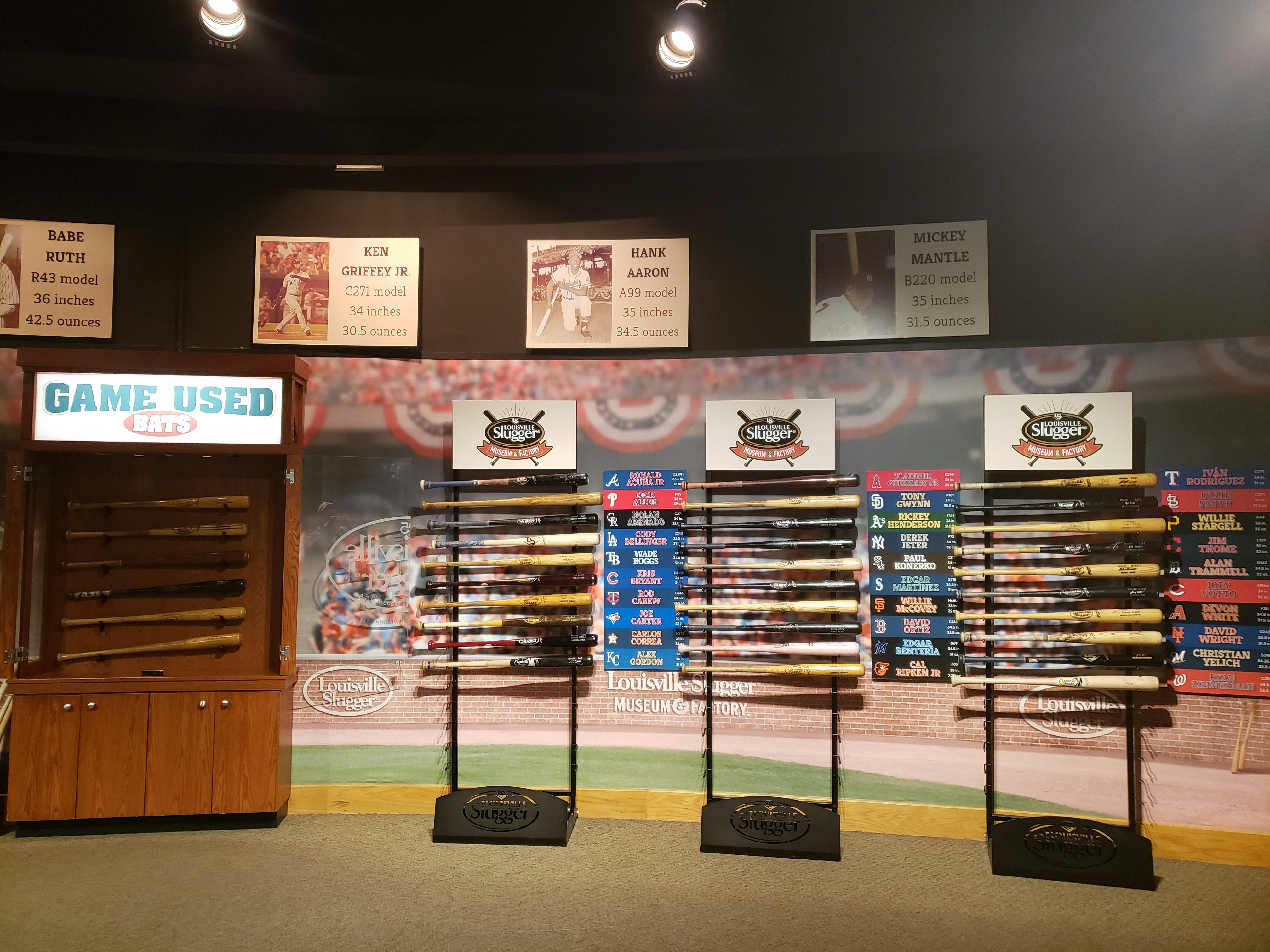 An interior shot of the Louisville Slugger Museum shows an exhibit of several bats used in baseball games by famous players. The bats are held on a special rack with the players' names next to each one. Overhead, signs show statistics for the size and weight of bats used by Babe Ruth, Hank Aaron, and other greats. Behind the bats, a large photographic wall mural simulates the crowd in a baseball stadium.jpg