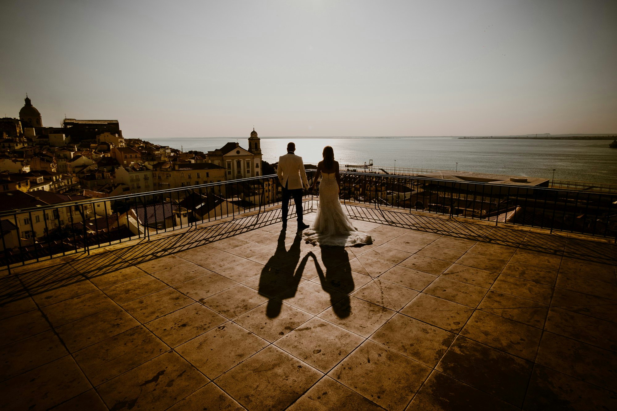 A bride and groom stand on a terrace with their backs to camera, gazing out over the sun setting on the sea.