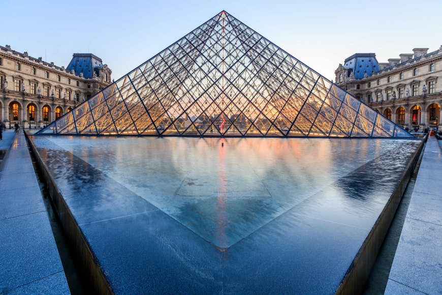 The glass pyramid of the Louvre at dusk, with its fountain in front and the ornate facades of the museum building on either side.