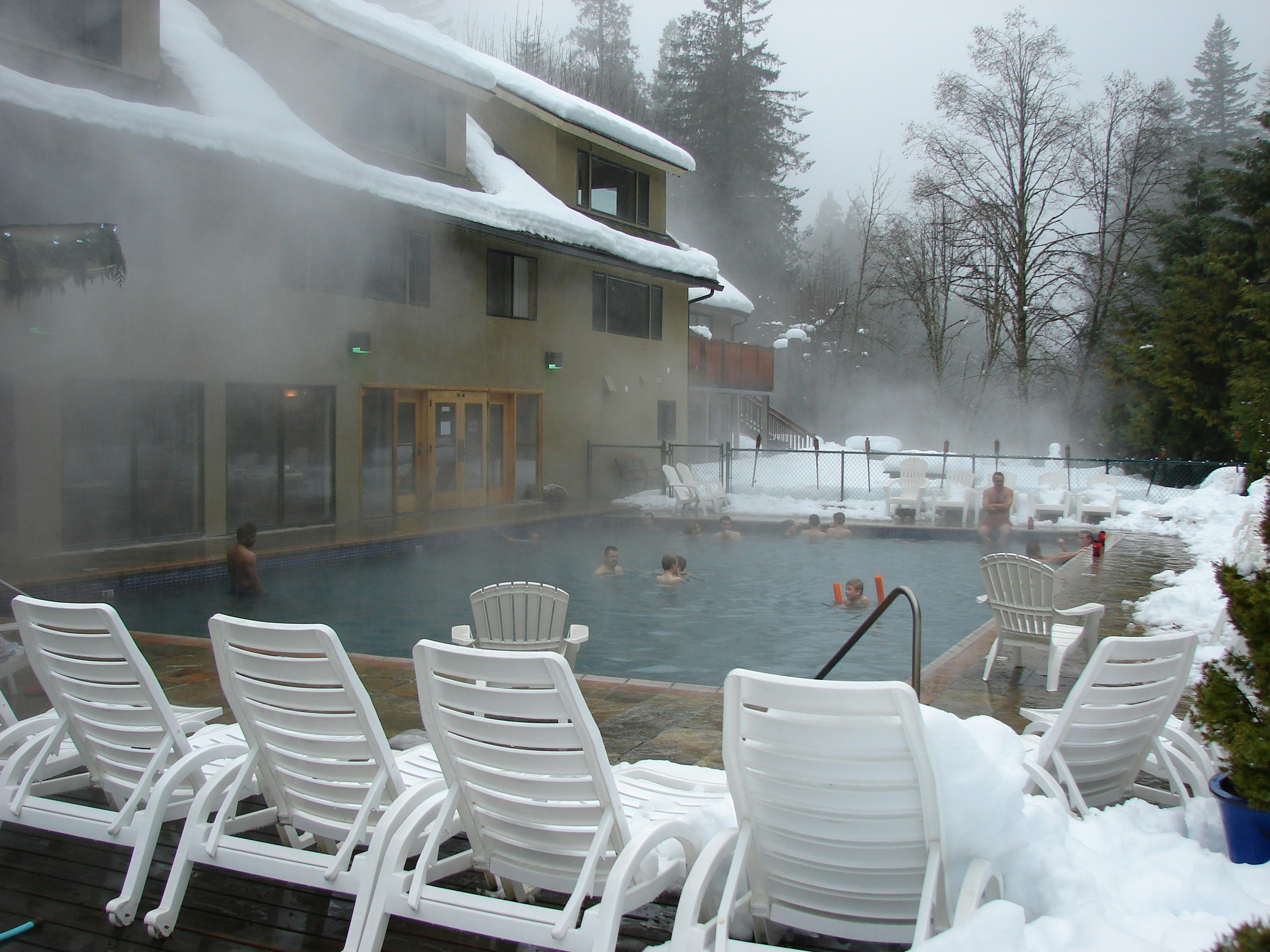 Steam rises against a grey sky and the snow-covered roof of Belknap Hot Springs Resort as bathers enjoy the blue waters of the pool, surrounded by plastic deck chairs as white as the snow.