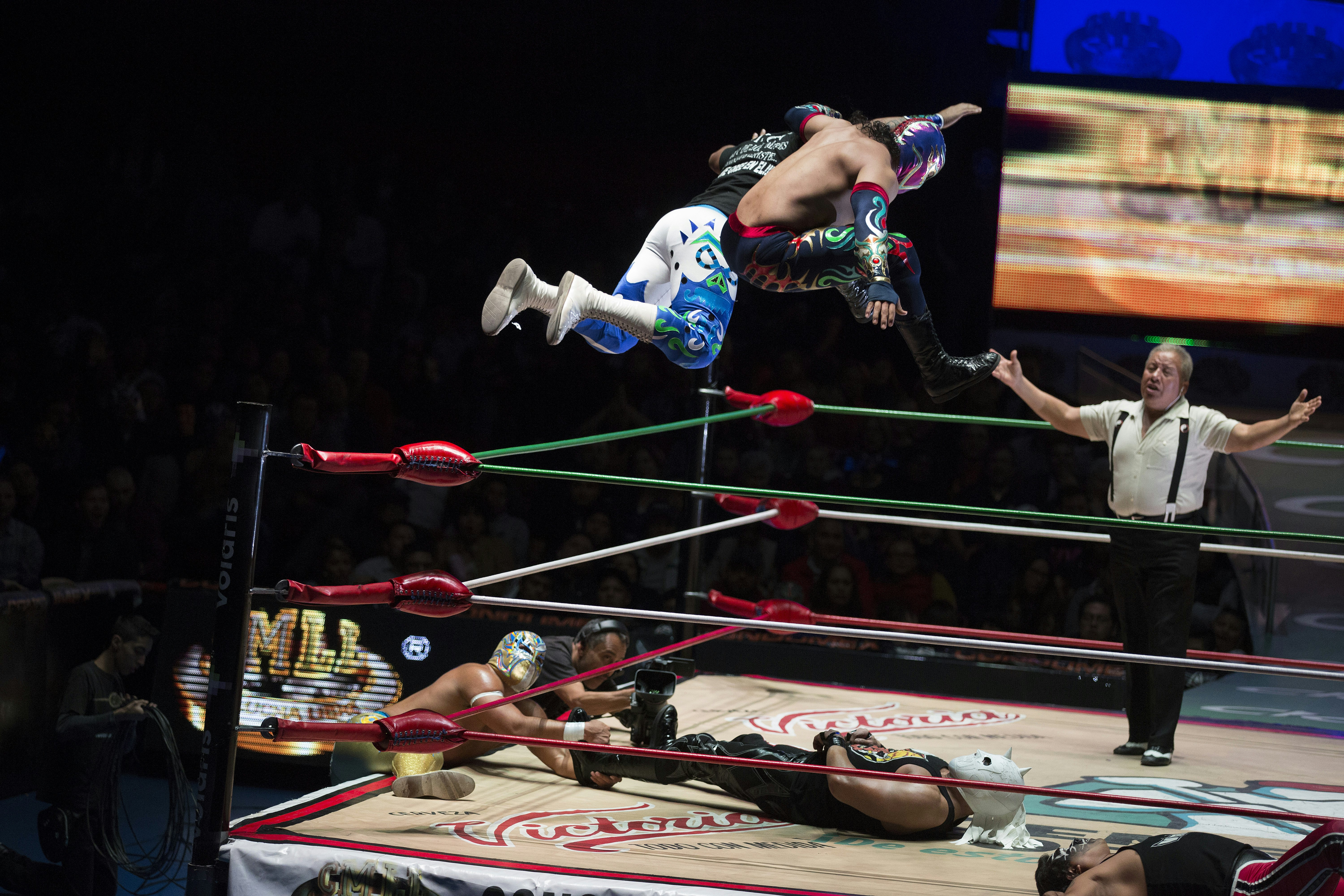 Dos luchadores salen volando del cinturón superior del ring de lucha mientras otro luchador se acuesta en la colchoneta.  Hay un árbitro con los brazos extendidos al fondo.