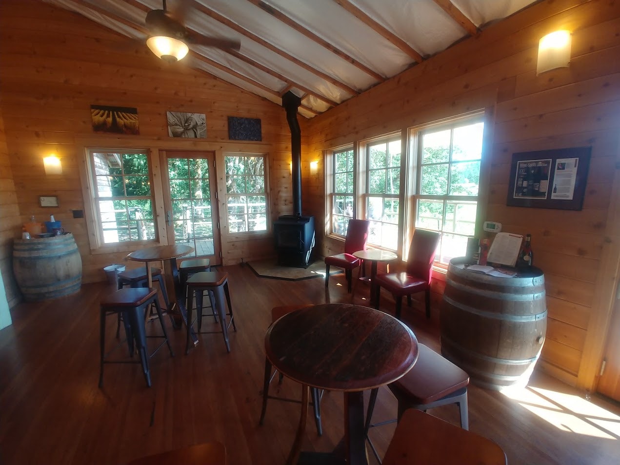 The warm and inviting interior of the Lumos Wine Company tasting room is made up on honey-wood plank walls and floor, with a cast iron wood stove in one corner and two high-top tables with pub stools in the room. Windows and a door look out on a deck around the building, while large wine barrels hold bottles and information booklets 