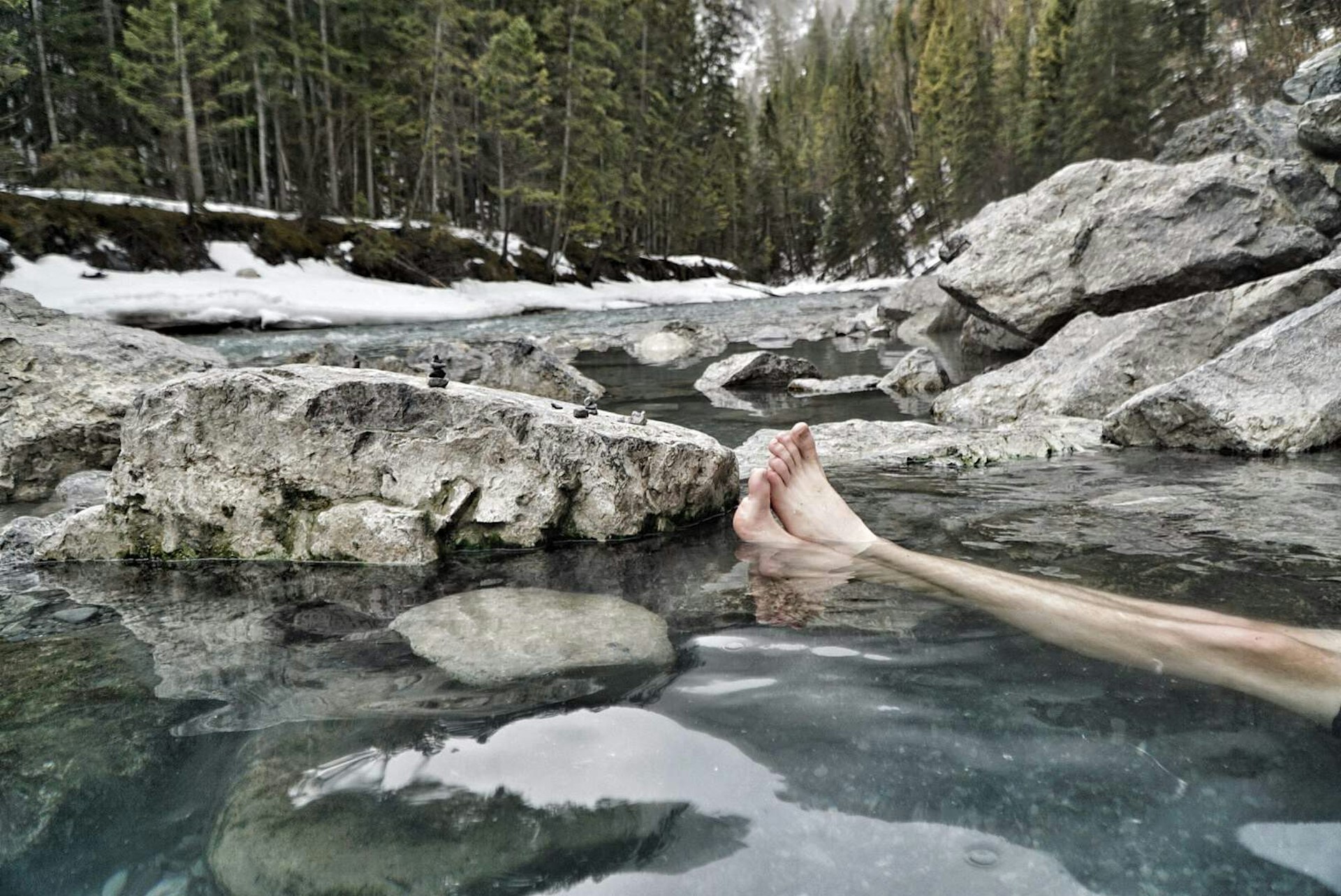 A person is sitting in a rocky pool, in the middle of a forest; only their crossed legs are visible.