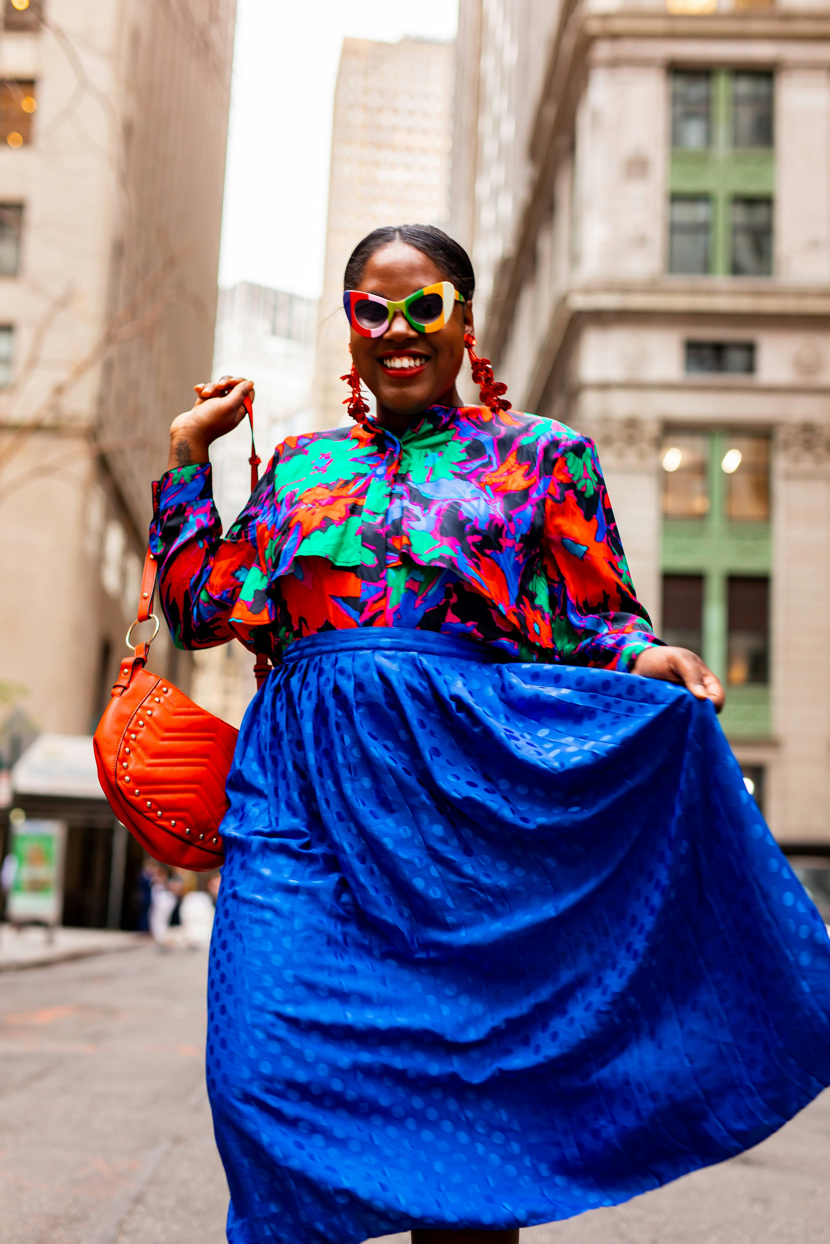 A smiling woman in flowered blouse and blue skirt smiles while carrying a red handbag